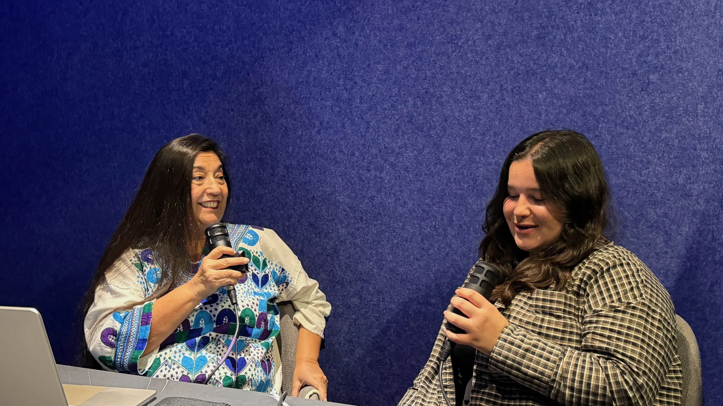 Two women sitting and talking into microphones in front of a blue wall, smiling and engaging in conversation.