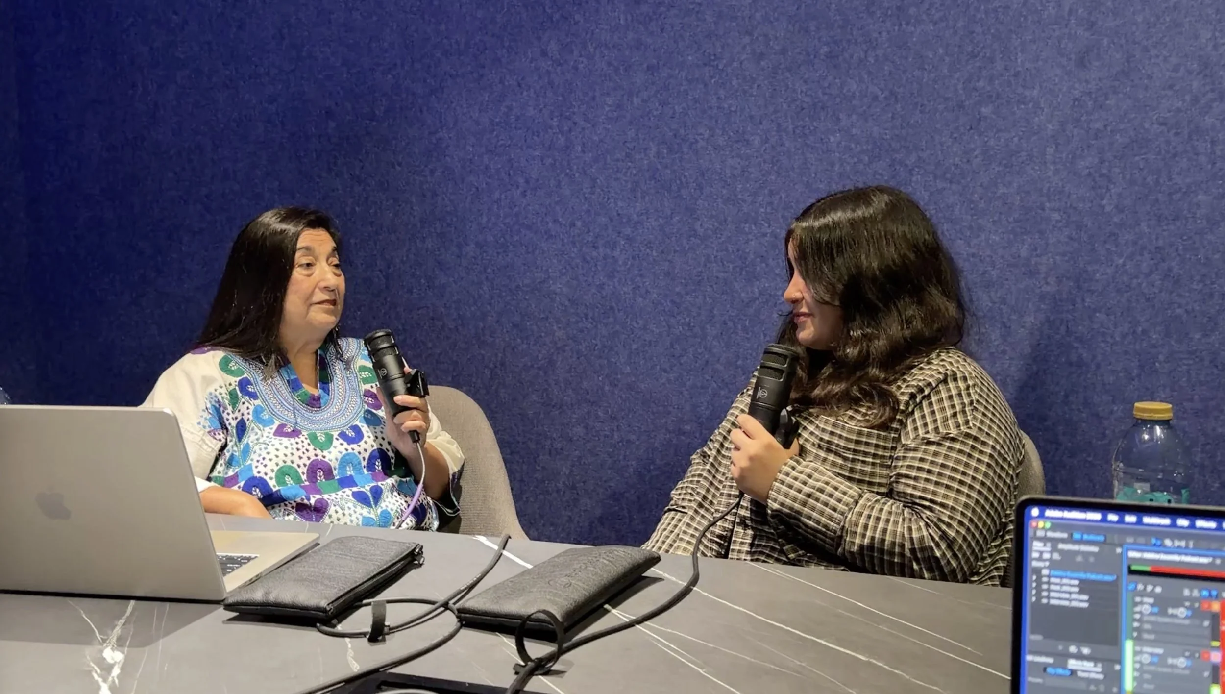 Two women sit at a table, each holding a microphone, engaged in a conversation. One woman wears a patterned blouse, and the other wears a checkered shirt. There are laptops, notebooks, and electronic equipment on the table, with a blue wall in the background.