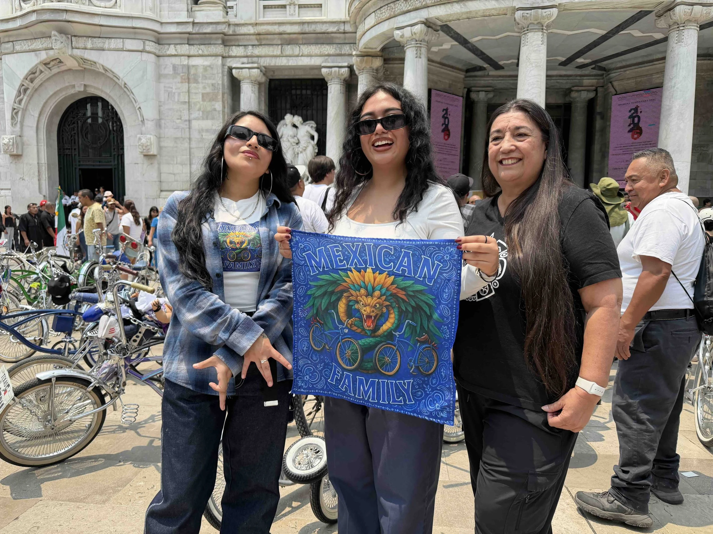 Three women standing together outdoors in front of a historic stone building with columns, holding a blue banner that reads 'Mexican Family', with bicycles in the background and a crowd of people.