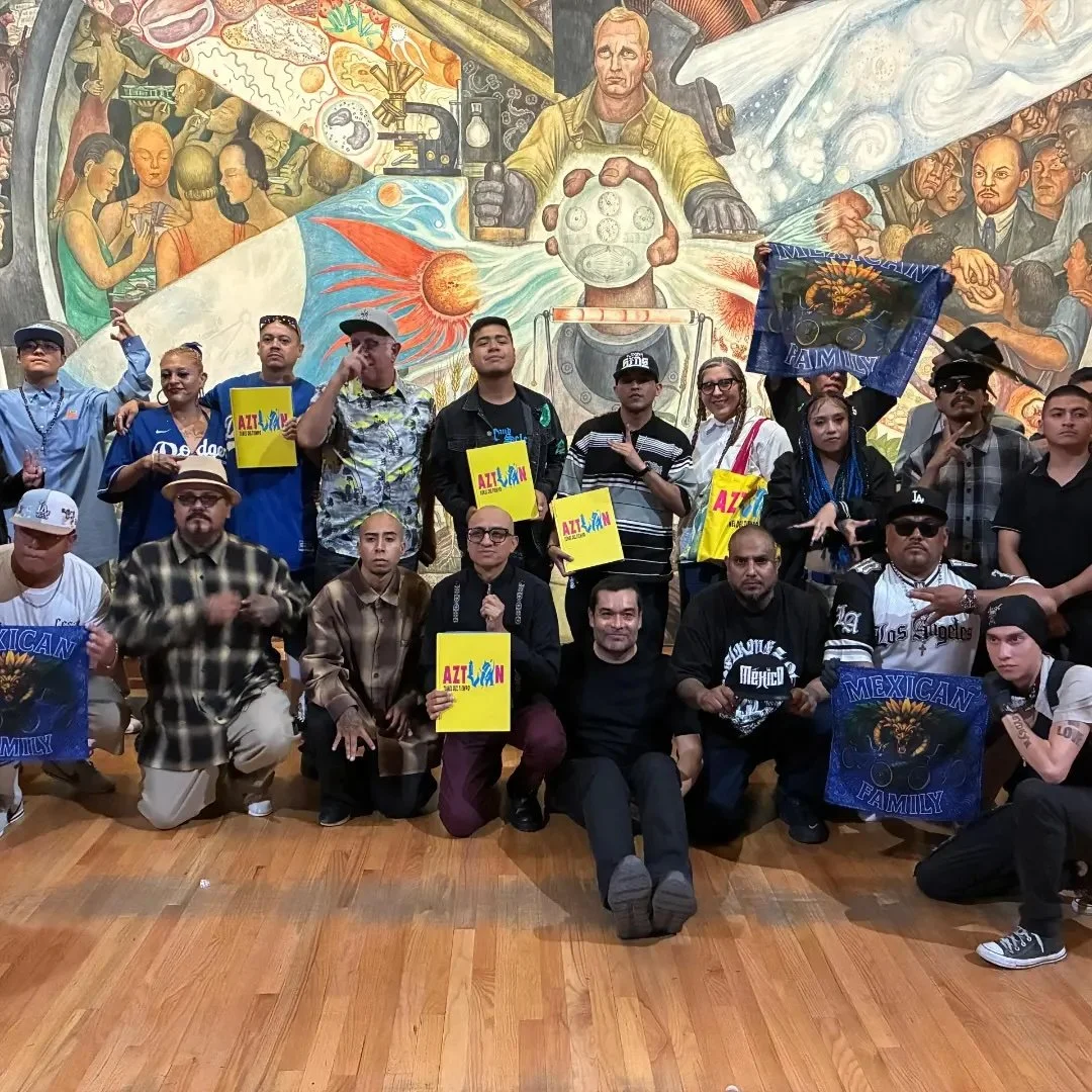 Group of people posing in front of a colorful mural, some holding signs and flags with the words "Mexican Family" and "AZTLAN".