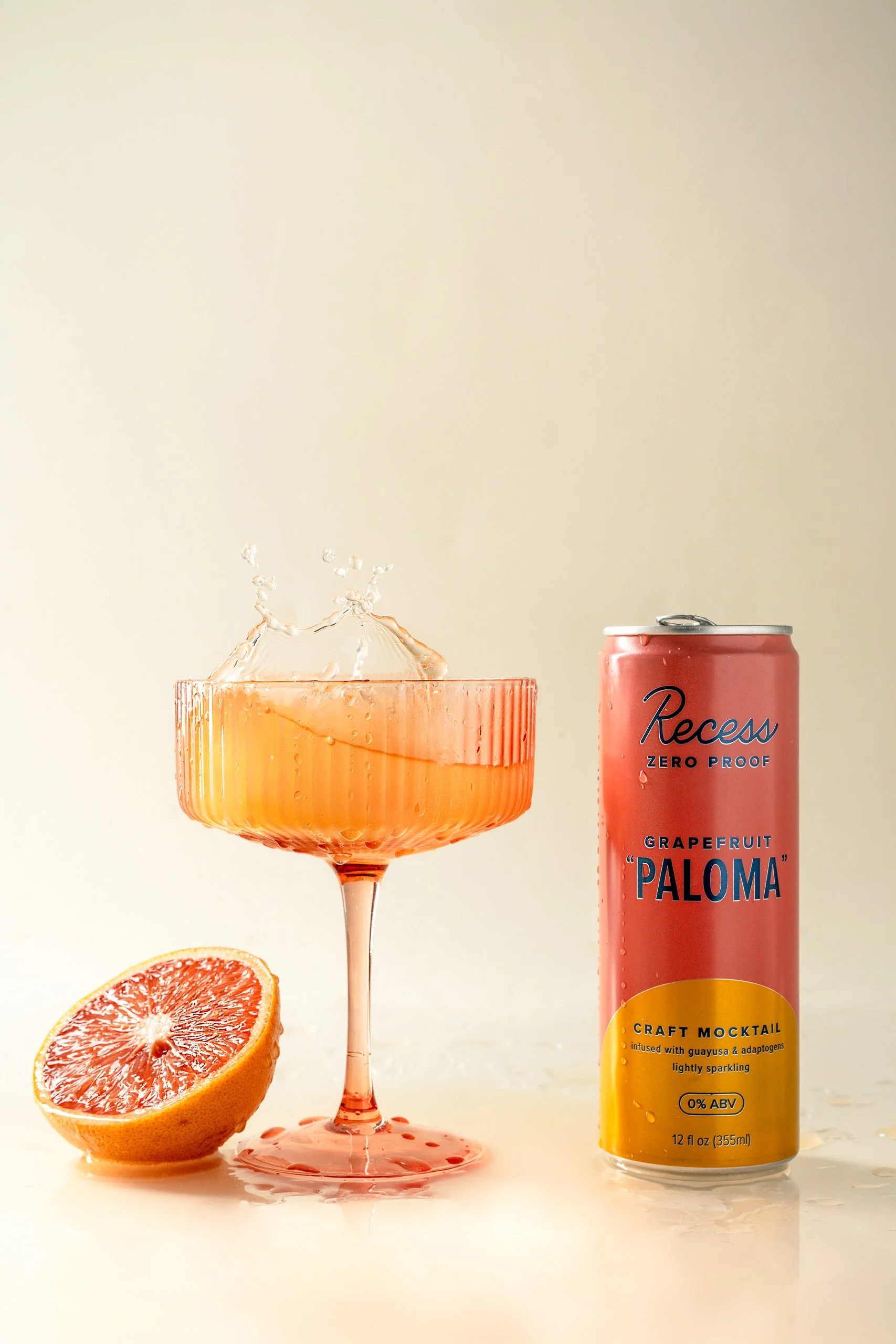 A sliced grapefruit, a pink grapefruit-flavored canned beverage called Paloma, and a glass with grapefruit juice and splash effects are arranged on a white surface against a beige background. The can describes the drink as a craft mocktail infused wi