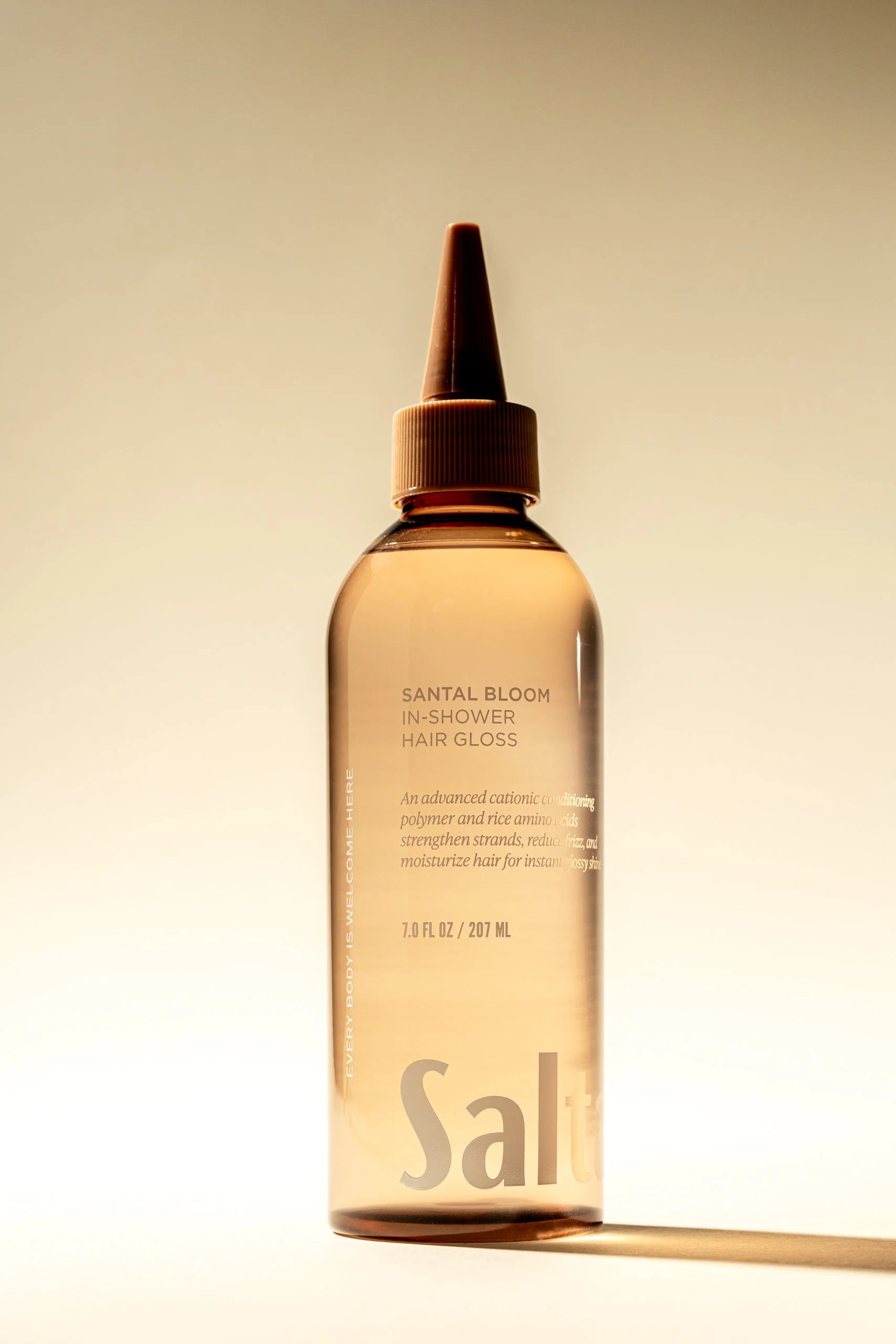 A clear plastic bottle of Santal Bloom in-shower hair gloss with a tan-colored conical nozzle. The label includes text about nourishing ingredients, set against a softly gradient background.