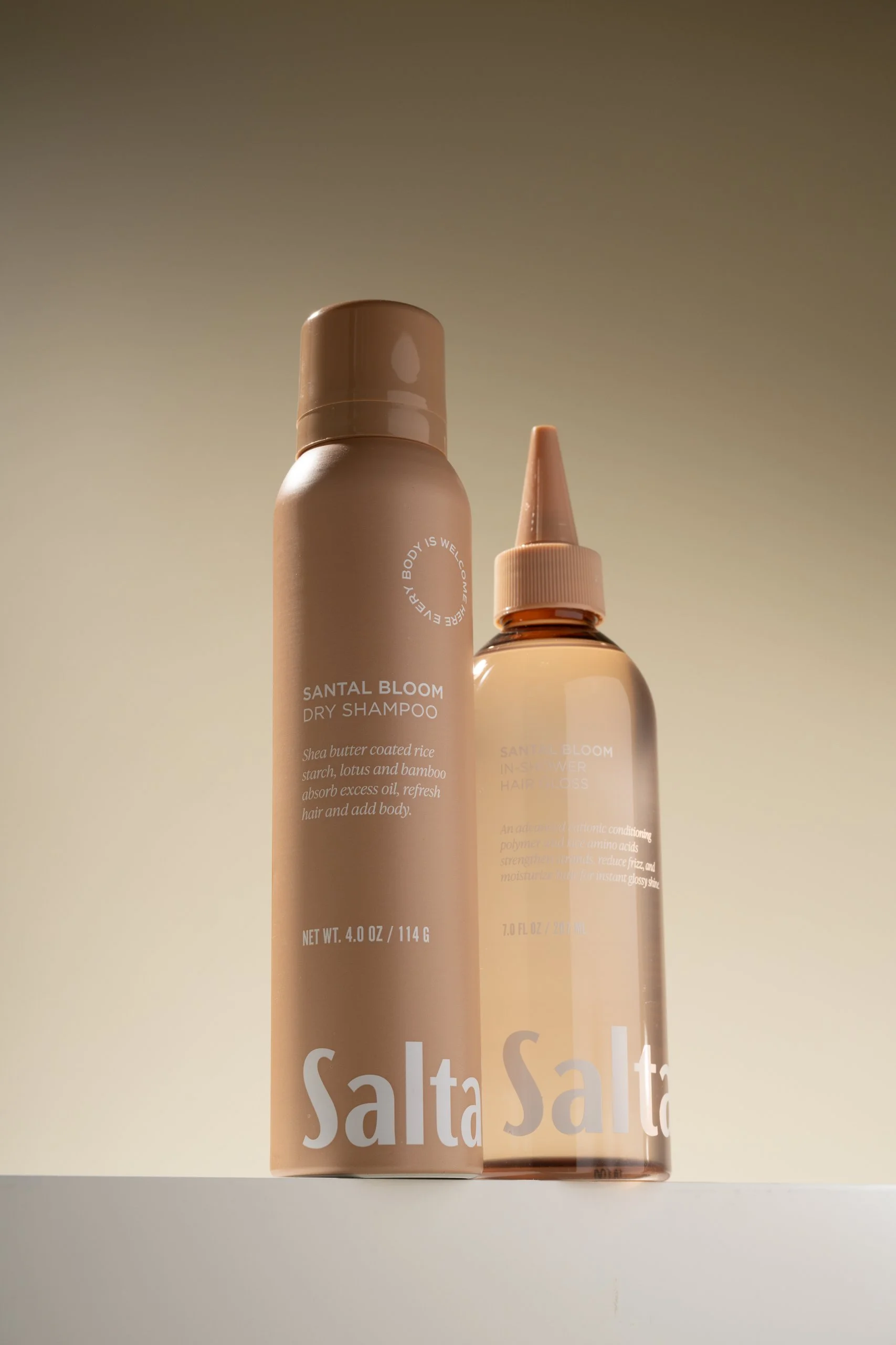 Two bottles of Saltair hair products, one labeled dry shampoo and the other hair gloss, with beige and brown packaging, against a plain background.