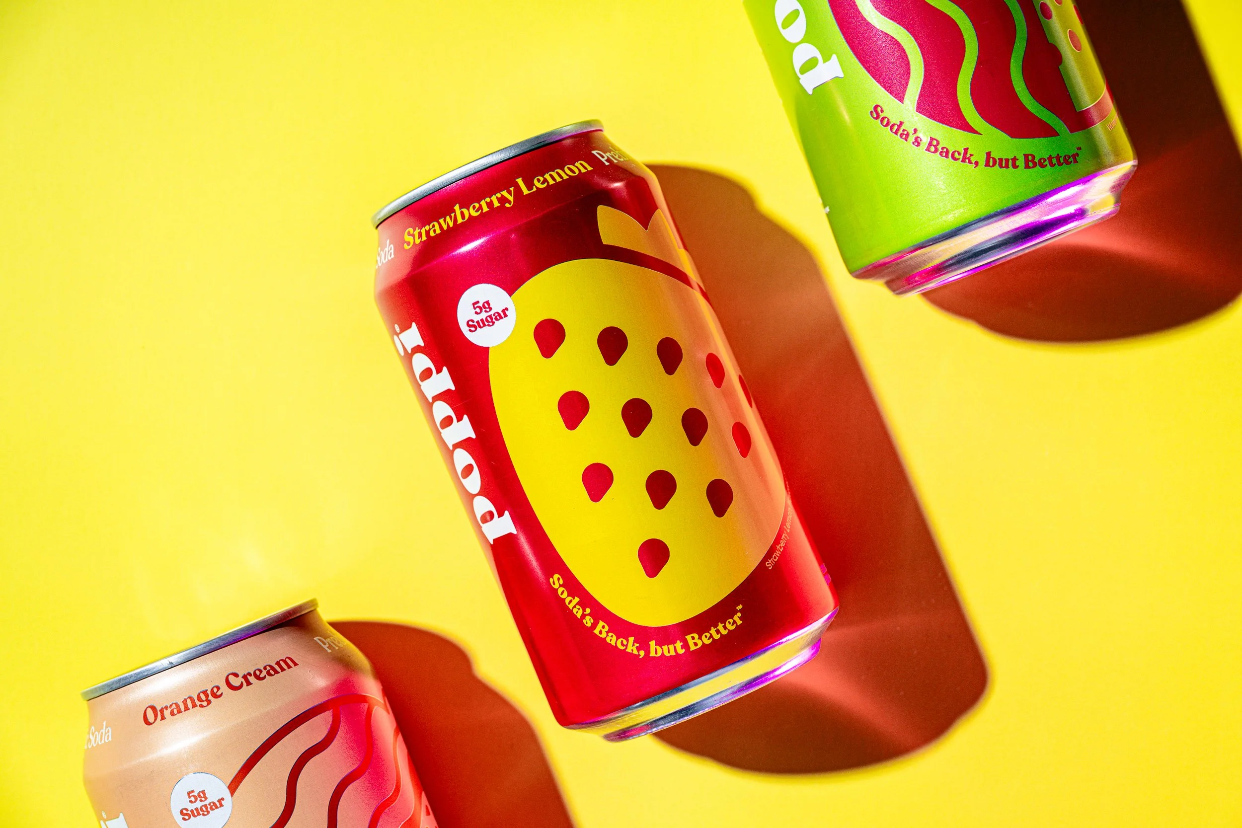 Cans of soda with colorful packaging featuring strawberry and orange flavors, placed on a bright yellow background.