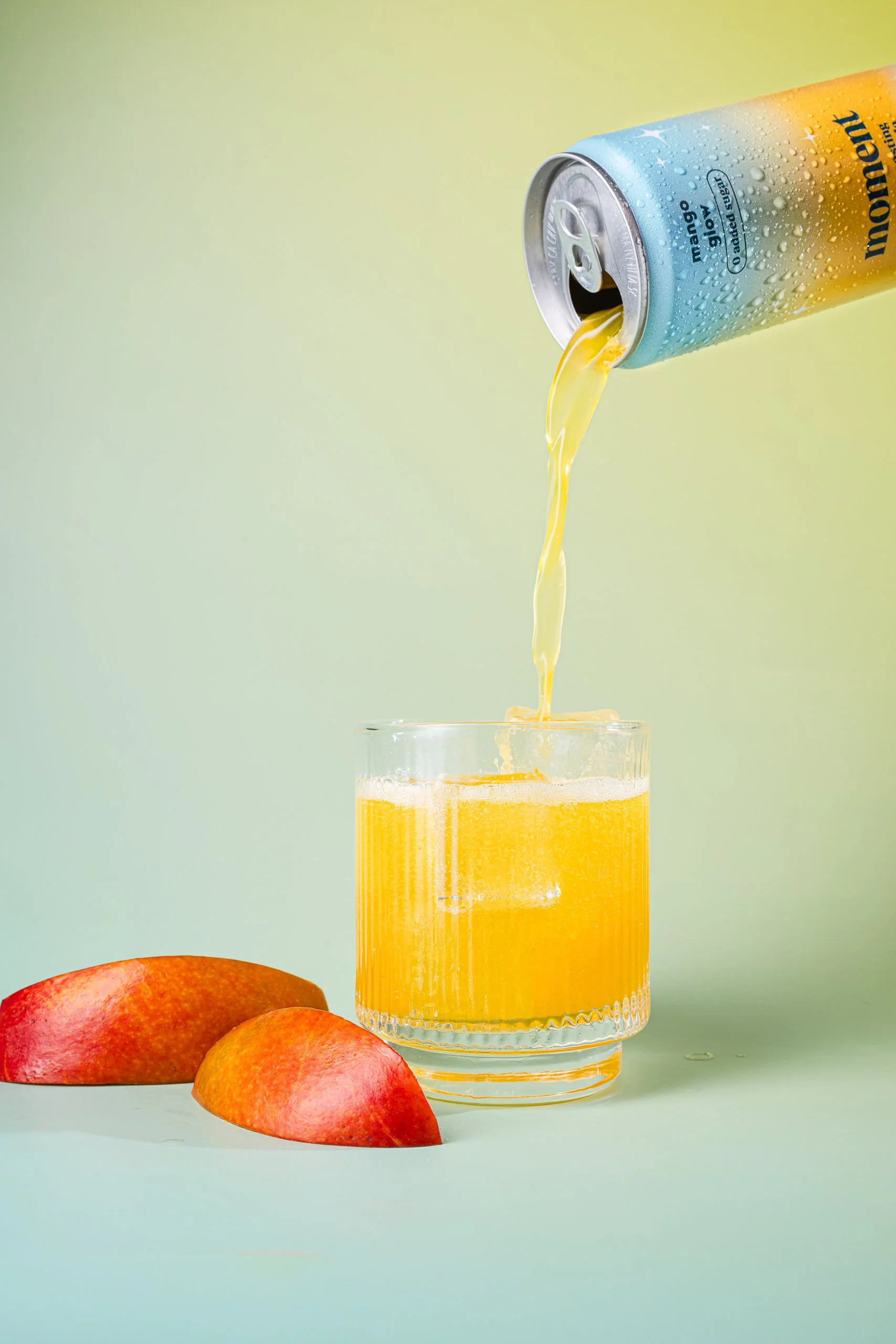 A can of mango-flavored sparkling water being poured into a glass, with three mangoes nearby on a light green background.