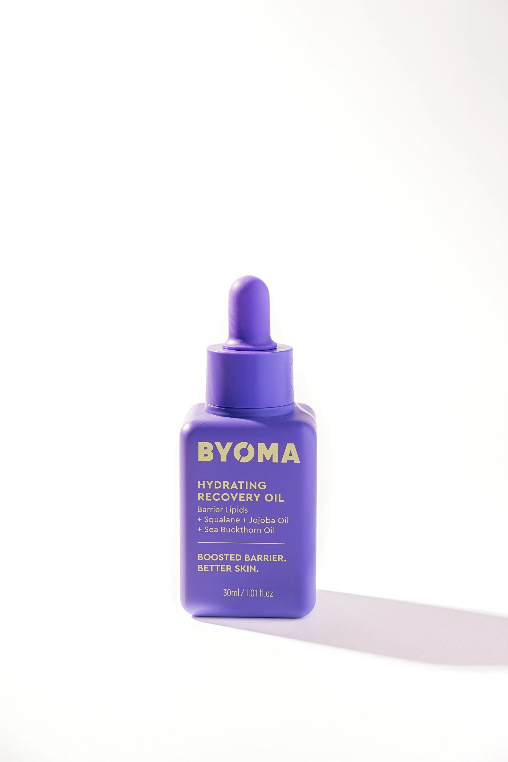 Purple bottle of BYOMA Hydrating Recovery Oil with a dropper cap, standing on a white surface. The label highlights barrier lipids, squalane, jojoba oil, and sea buckthorn oil.