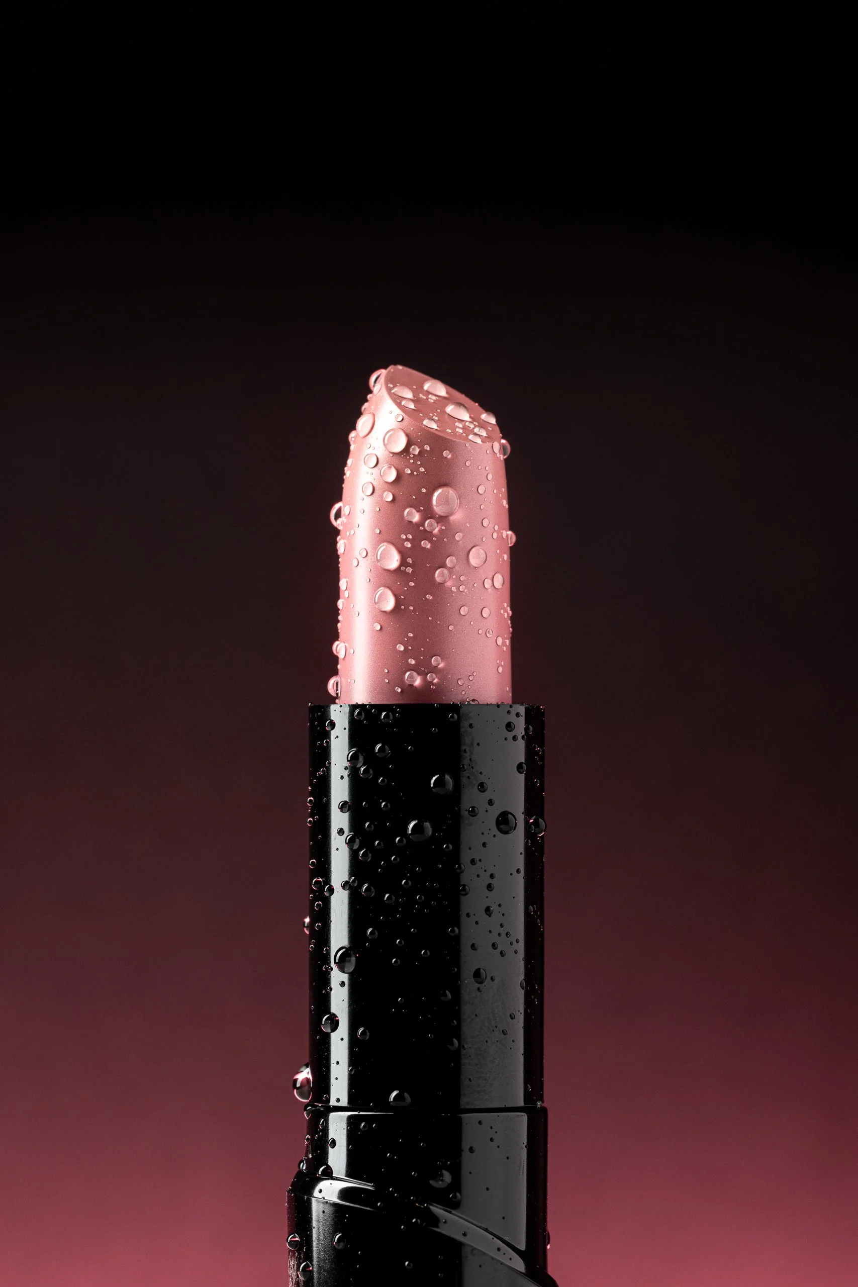 Close-up of a pink lipstick with water droplets on its surface, set against a dark background.