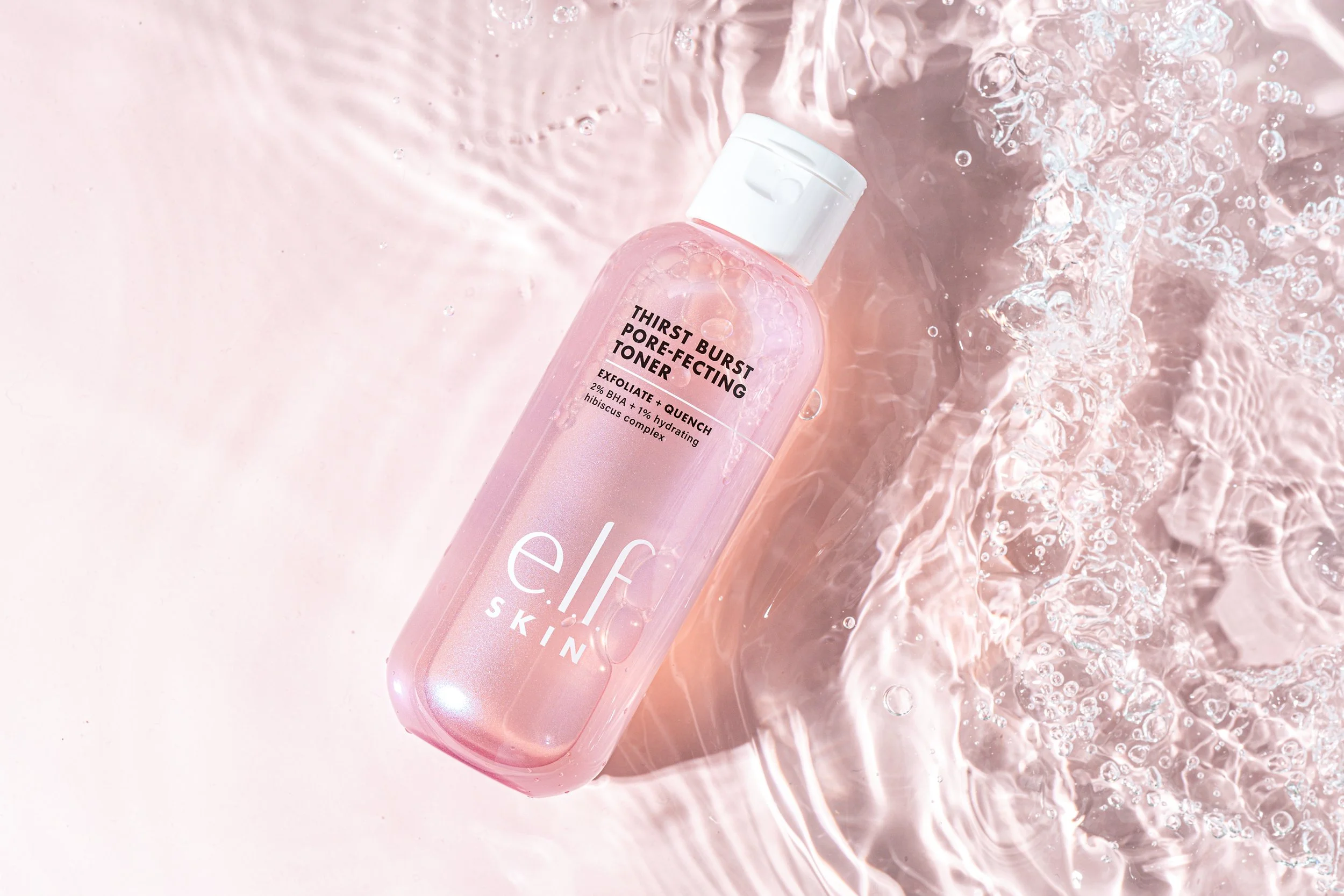 Product photography for skincare brand with clean studio lighting. A pink bottle of skincare toner labeled "Thirst Burst Pore-Refining Toner" floating in water with bubbles and ripples.