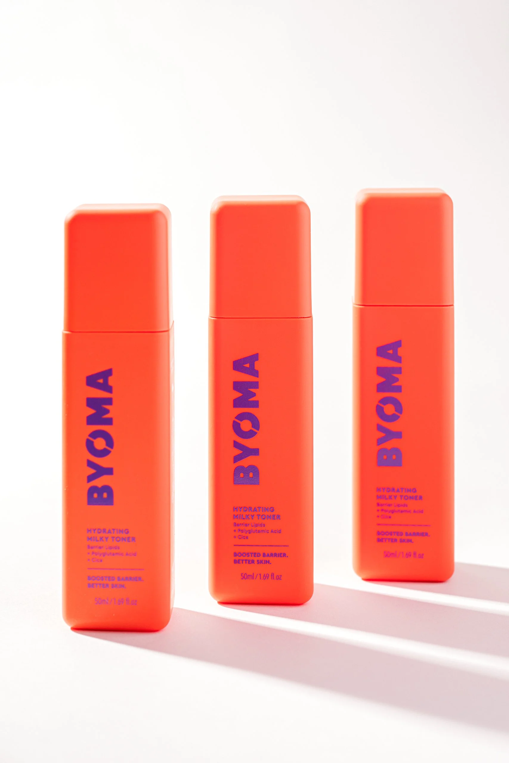 Three orange bottles of BYOMA Hydrating Milky Toner with blue text on a white background, casting soft shadows.