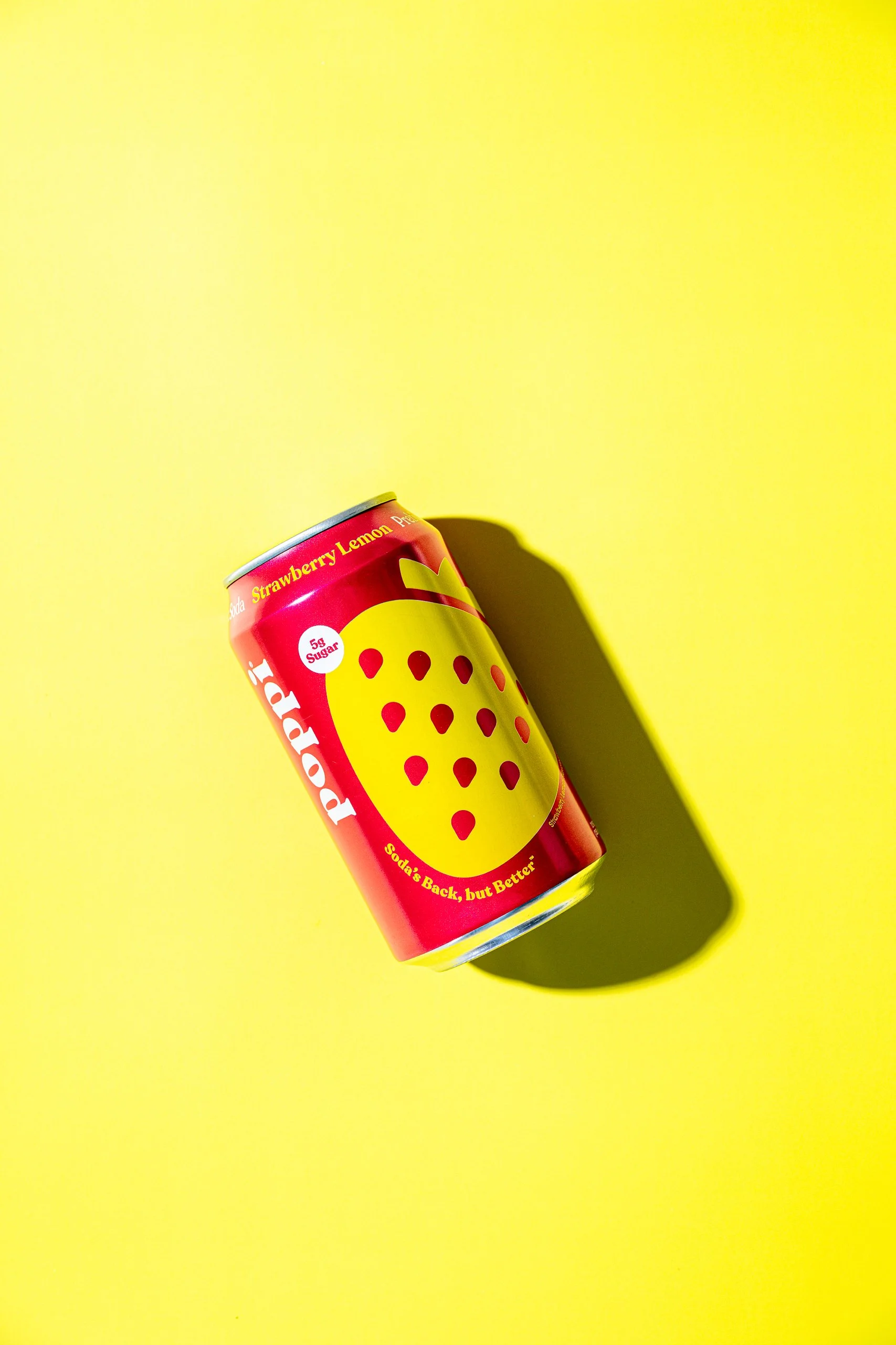 A red soda can with a yellow label featuring a strawberry illustration, set against a bright yellow background, casting a shadow.