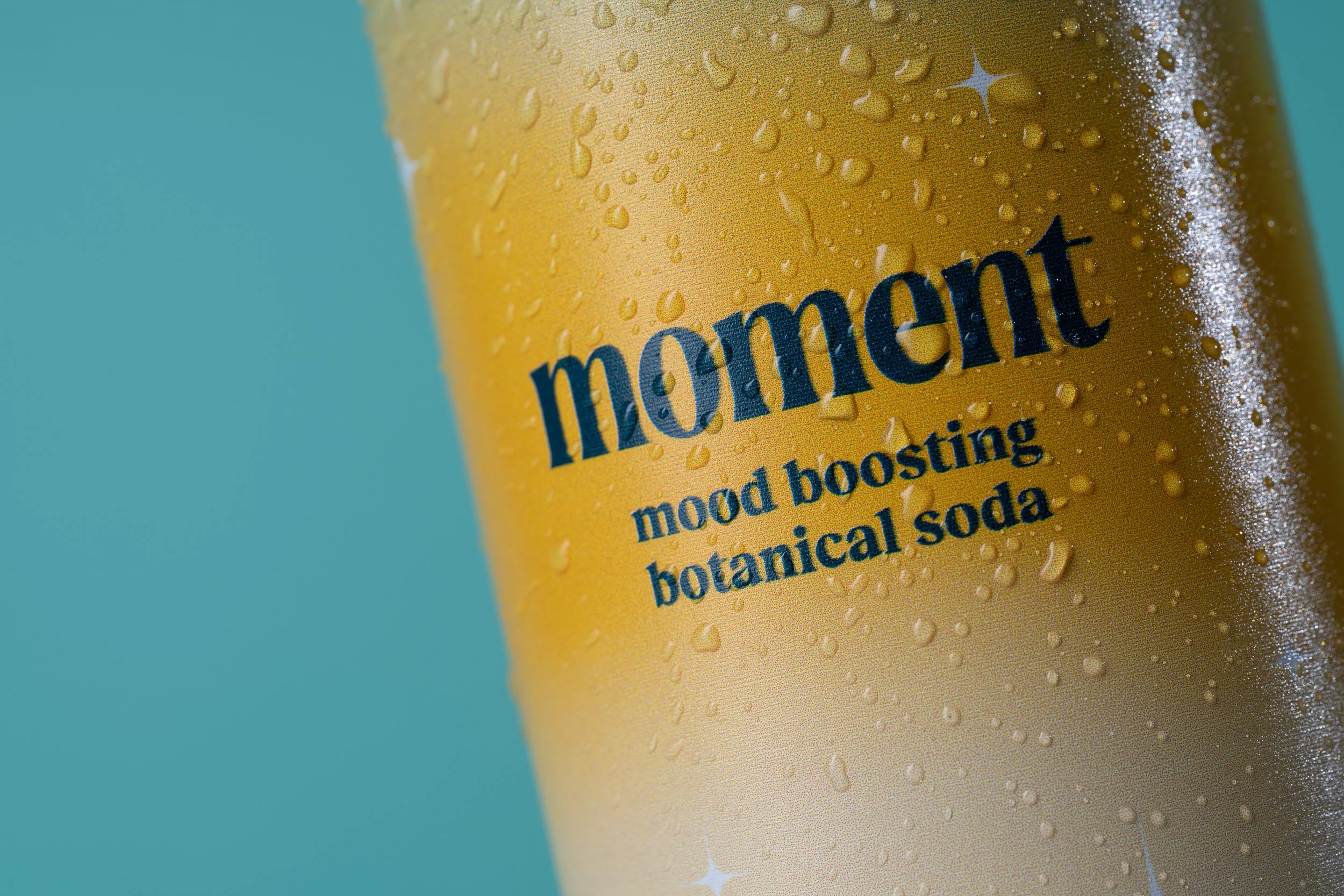 Close-up of a yellow beverage can with condensation droplets, featuring the word 'moment' and the phrase 'mood boosting botanical soda' on the label, against a light blue background.