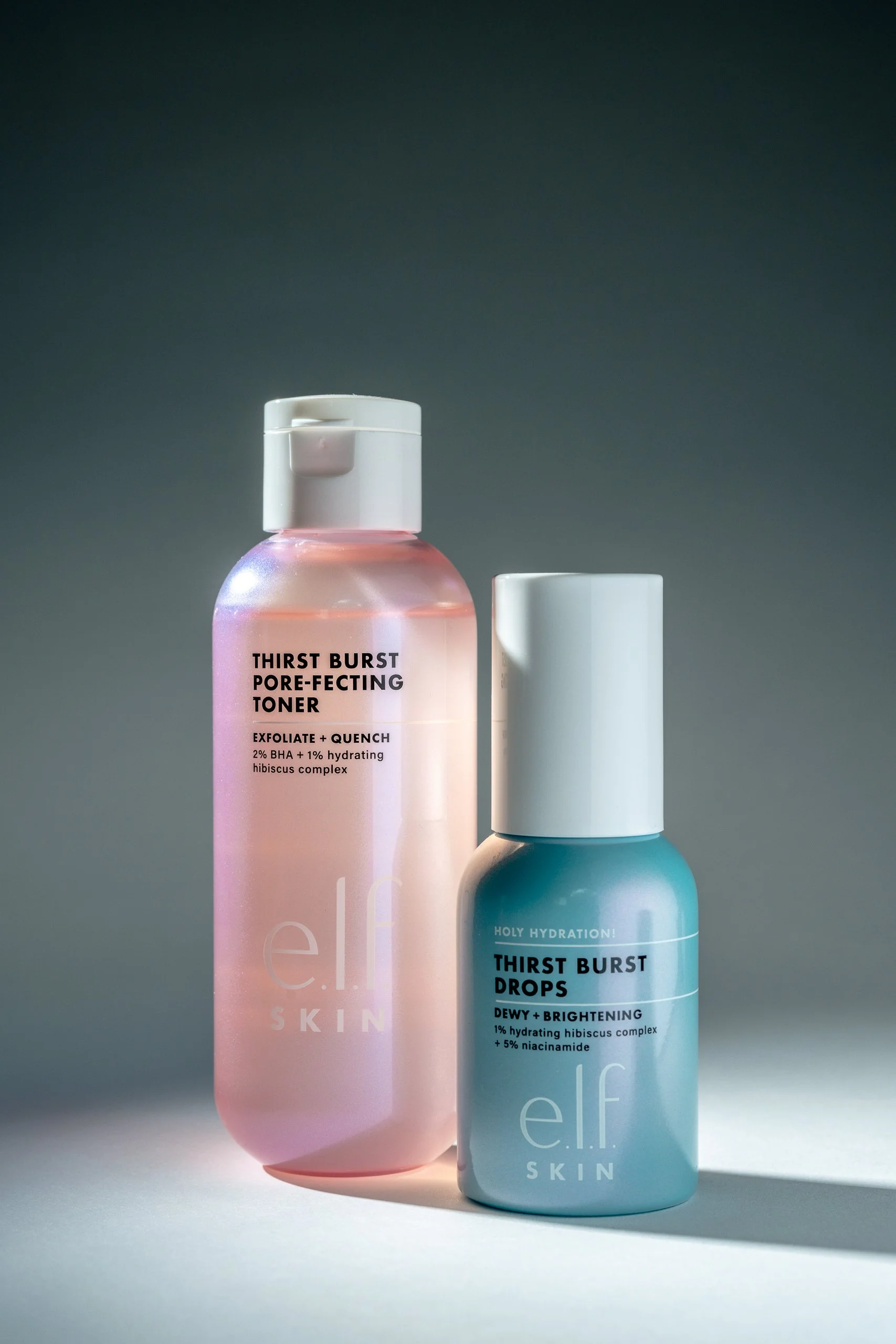 Two skincare bottles from the brand e.l.f. Skin with pastel-colored packaging; the pink bottle is labeled 'Thirst Burst Pore-Perfecting Toner' and the blue bottle is labeled 'Thirst Burst Drops.' Both bottles have white caps and are set against a gradient background.