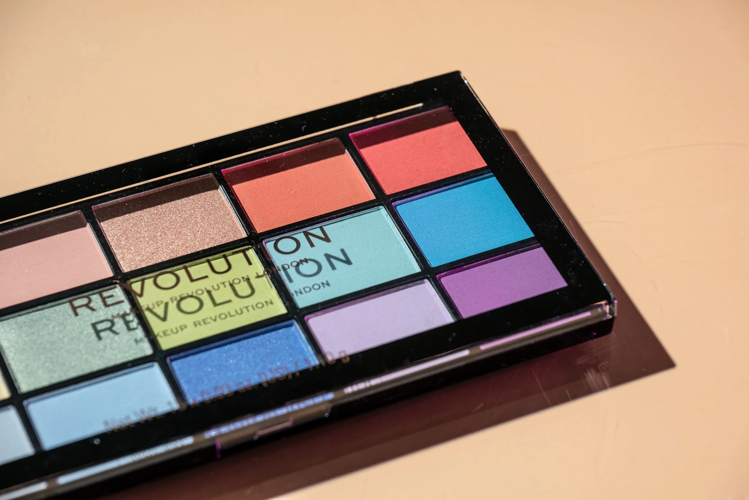 Close-up of a colorful eyeshadow palette with multiple shades including pink, blue, purple, and beige arranged in a grid, with the text 'REVOLUTION' repeated on some of the pans.