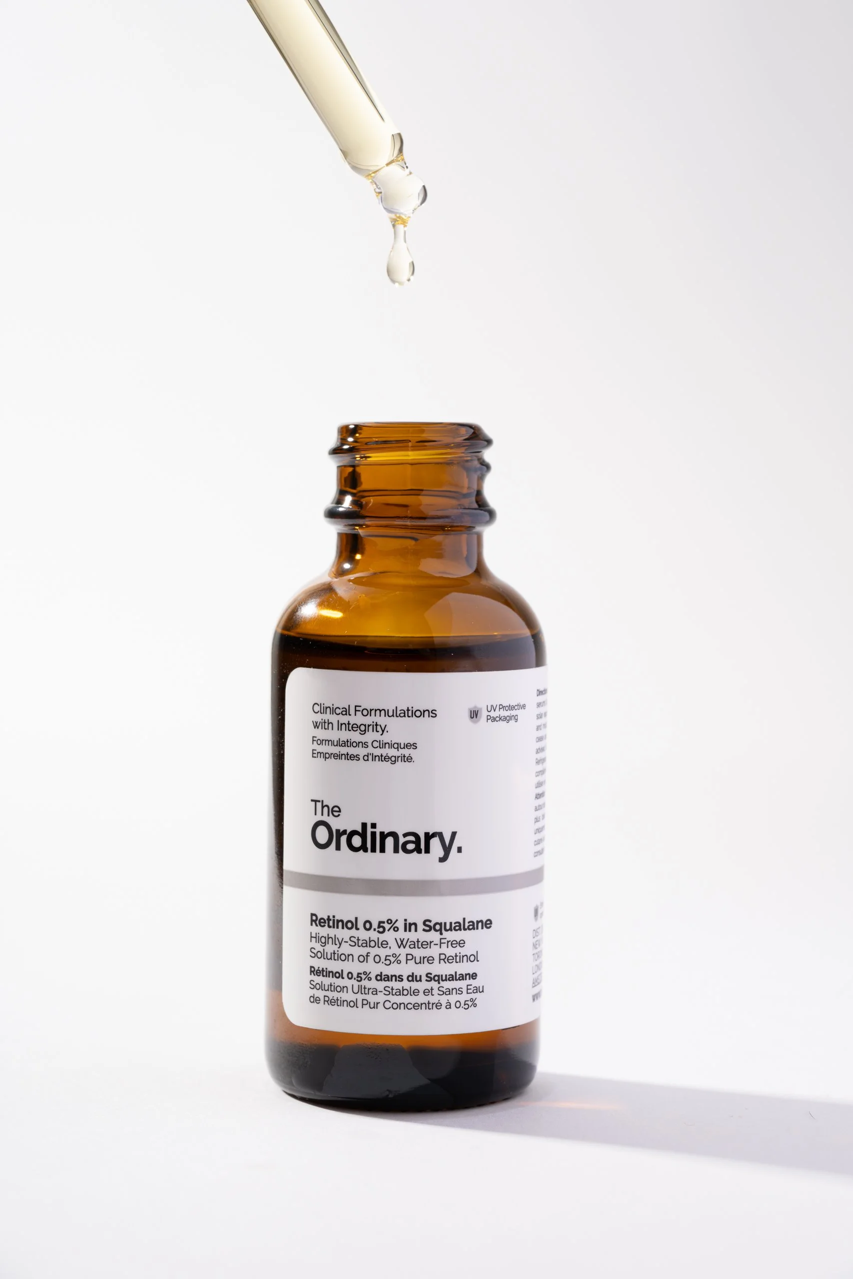 A brown glass bottle of The Ordinary Retinol 0.5% in Squalane with a label that displays product information, and a pipette releasing a droplet of liquid into the bottle, set against a plain white background.