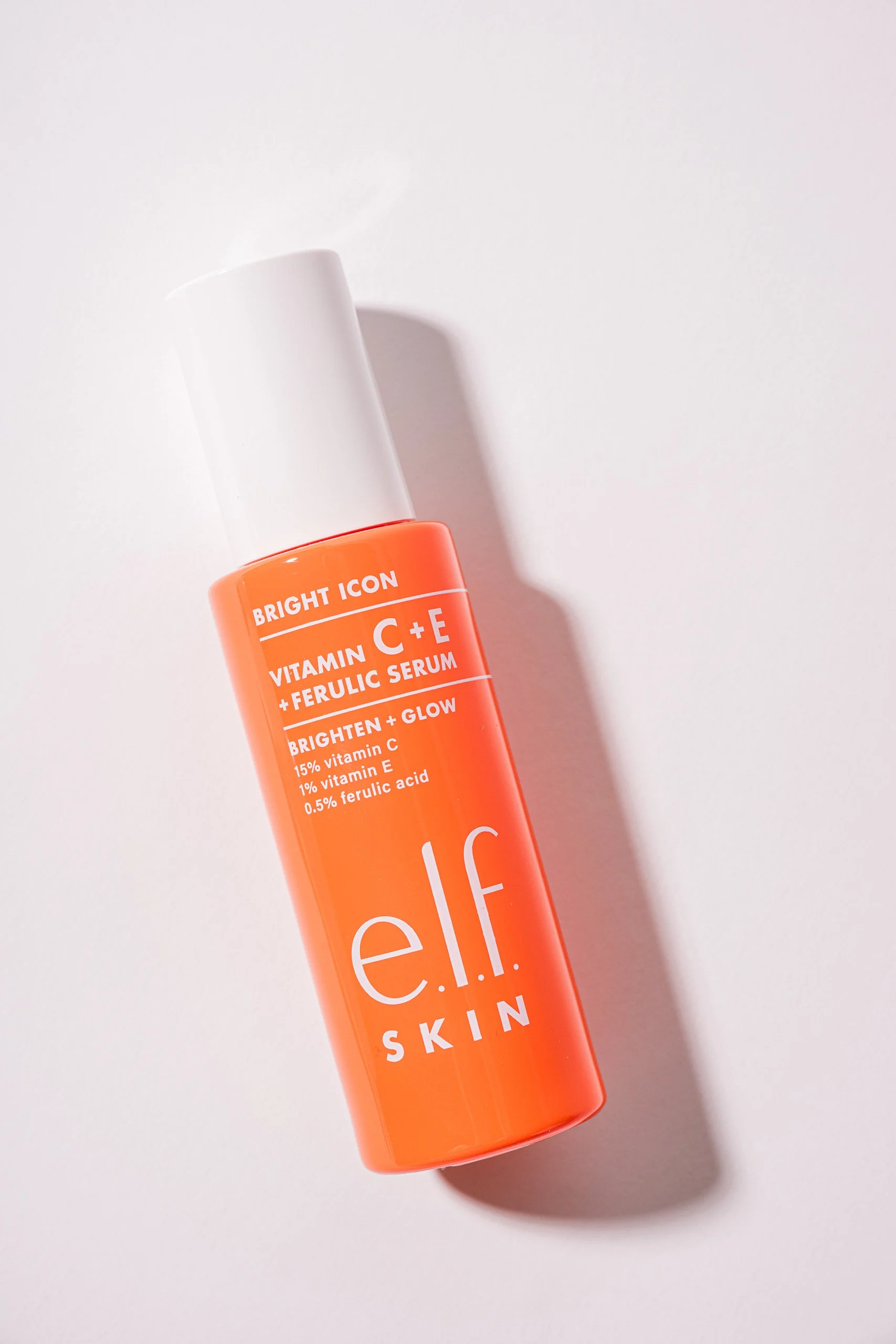 Close-up of a bright orange bottle of skincare serum with white text, against a white background, partially cropped, casting a shadow.