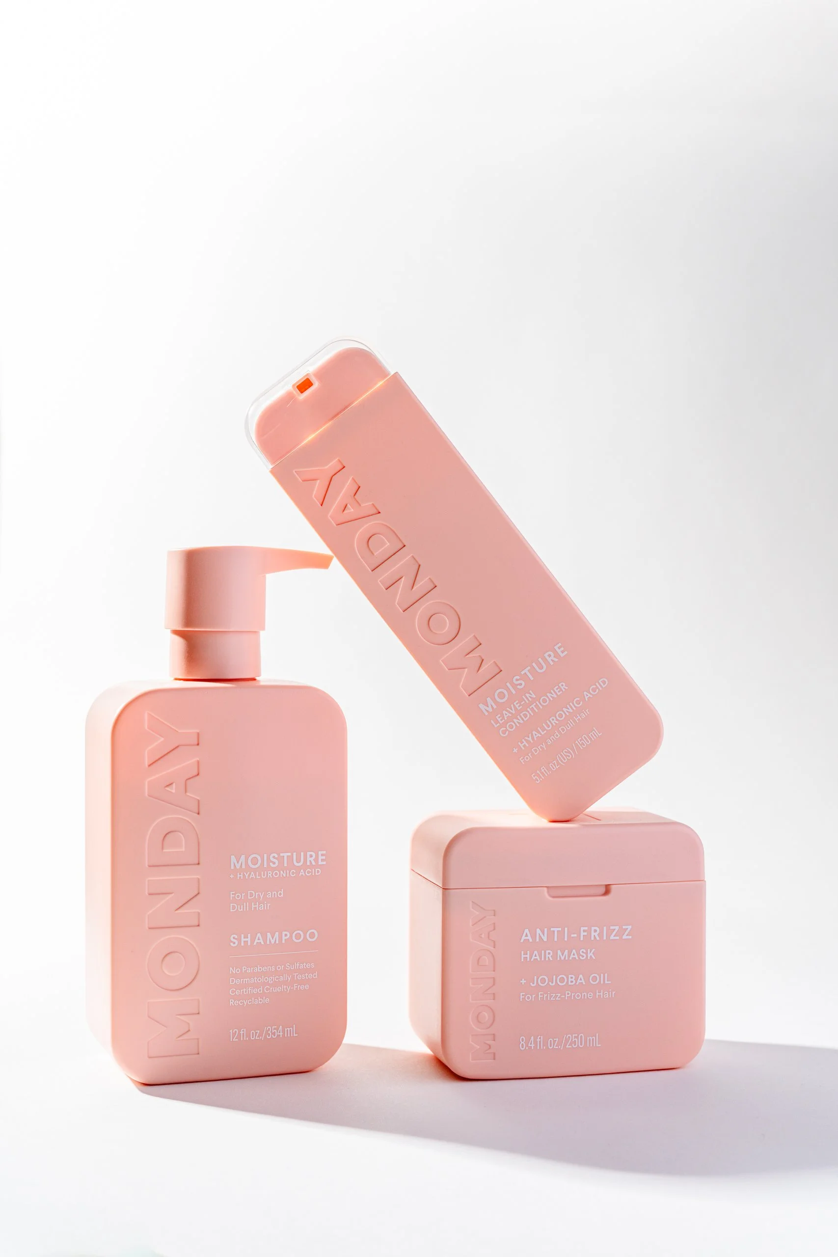Pink bottles of hair care products with minimalist packaging on white background, including shampoo, leave-in conditioner, and hair mask.