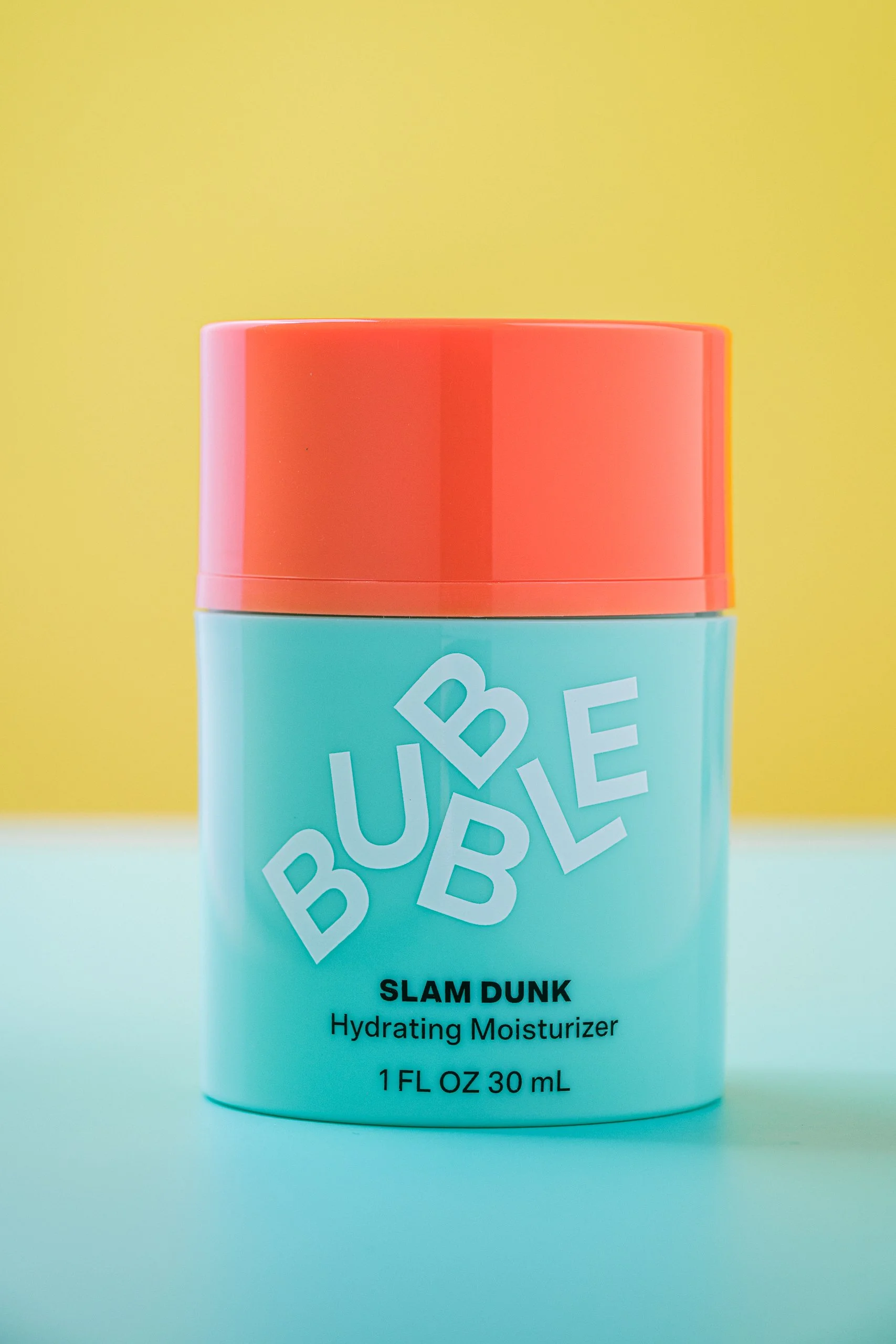 A cylindrical blue container with a coral-colored lid. The container features the label 'BUBBLE' in large white letters, and the text 'SLAM DUNK Hydrating Moisturizer 1 FL OZ 30 mL' in smaller black letters below. The background is a vibrant yellow.