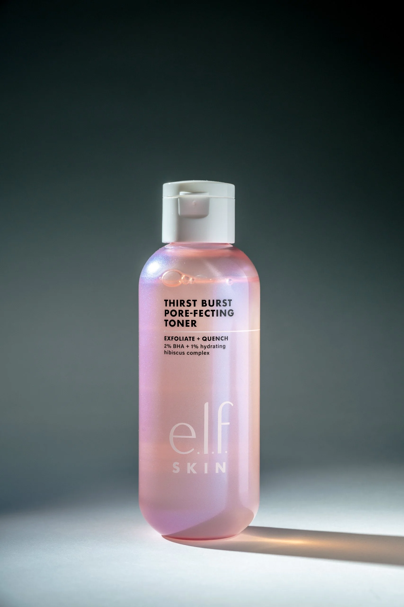 A pink bottle of e.l.f. skincare toner with a white flip-top cap, labeled 'Thirst Burst Pore-Refing Toner' with ingredients 2% BHA and 1% hydrating hibiscus complex, standing on a gray surface with a dark background.