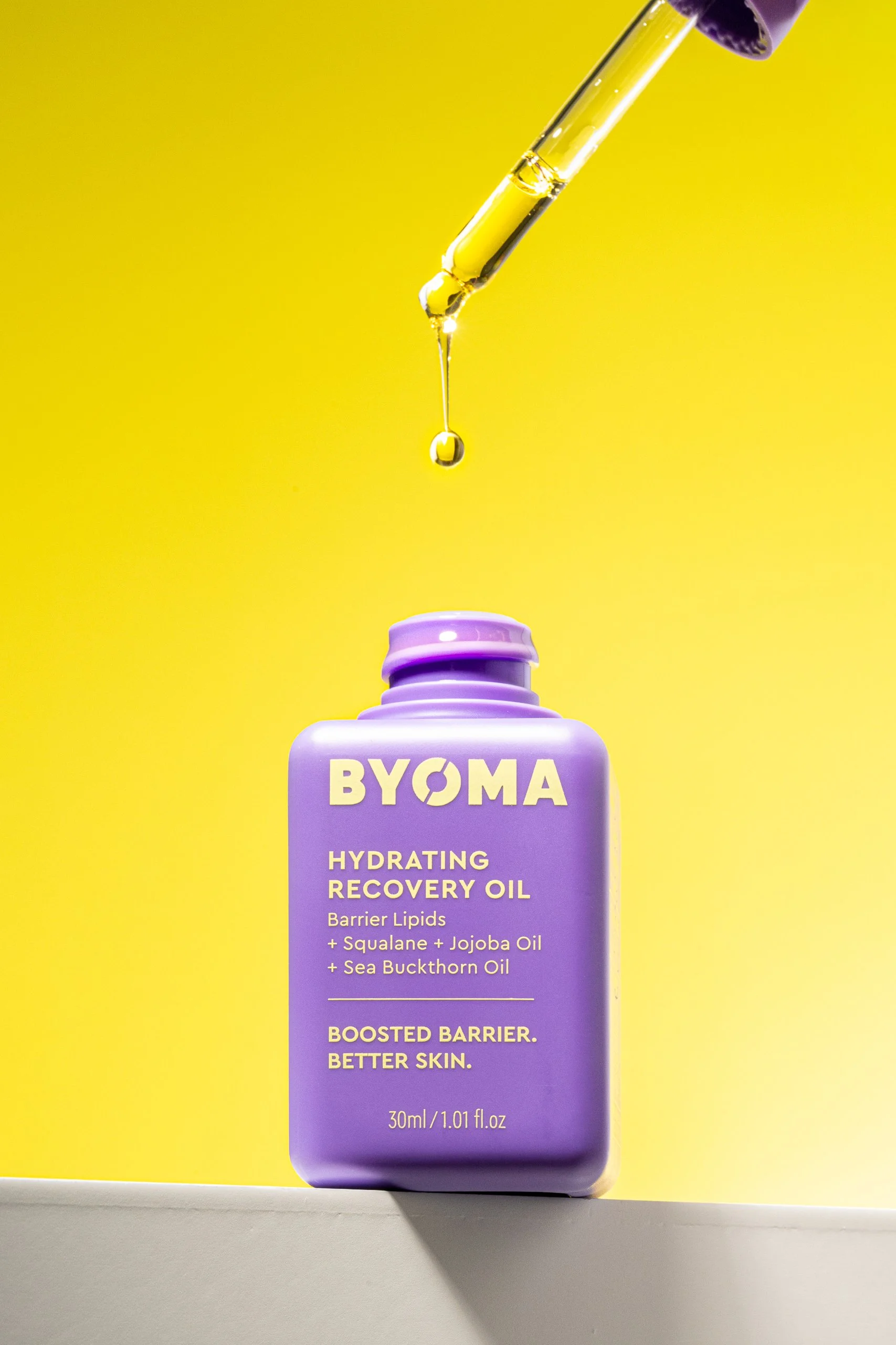 A purple bottle of BYOMA Hydrating Recovery Oil with a yellow background. The bottle has white text describing the product and ingredients, and a dropper dispensing a drop of oil into the air.