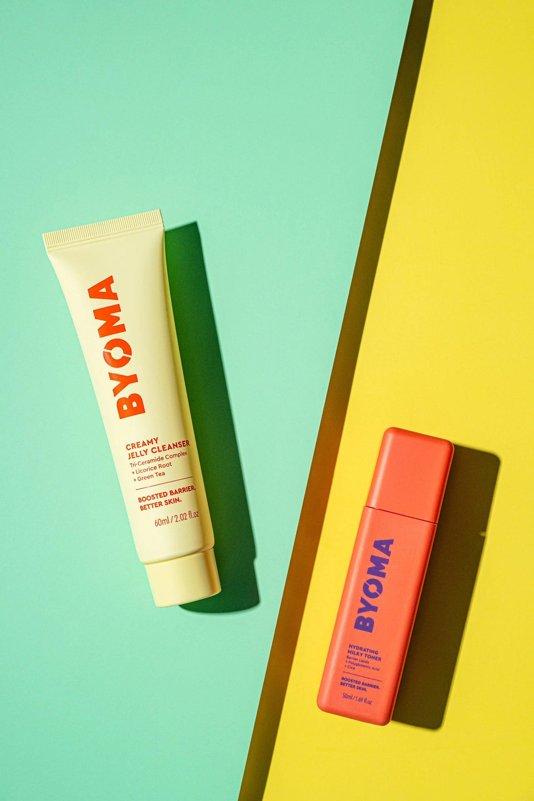 A flat lay of two skincare products, a yellow tube labeled 'Byoma' with red text and a pink bottle also labeled 'Byoma' with purple text, on a colorful background of mint green, yellow, and teal panels with shadows.