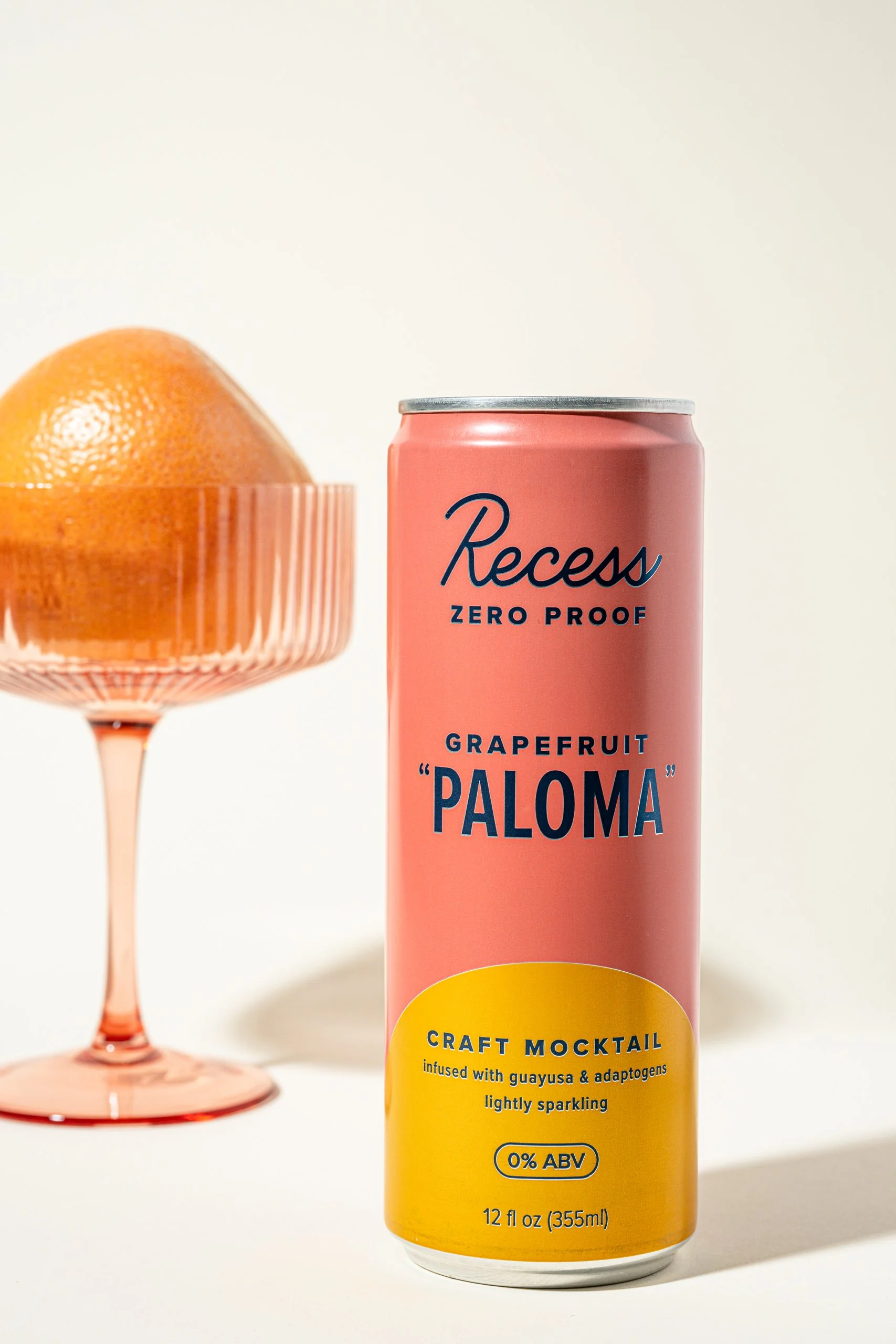 A pink and yellow can of Recess Grapefruit Paloma craft mocktail drink placed next to a pink glass with a grapefruit inside it, on a white background.