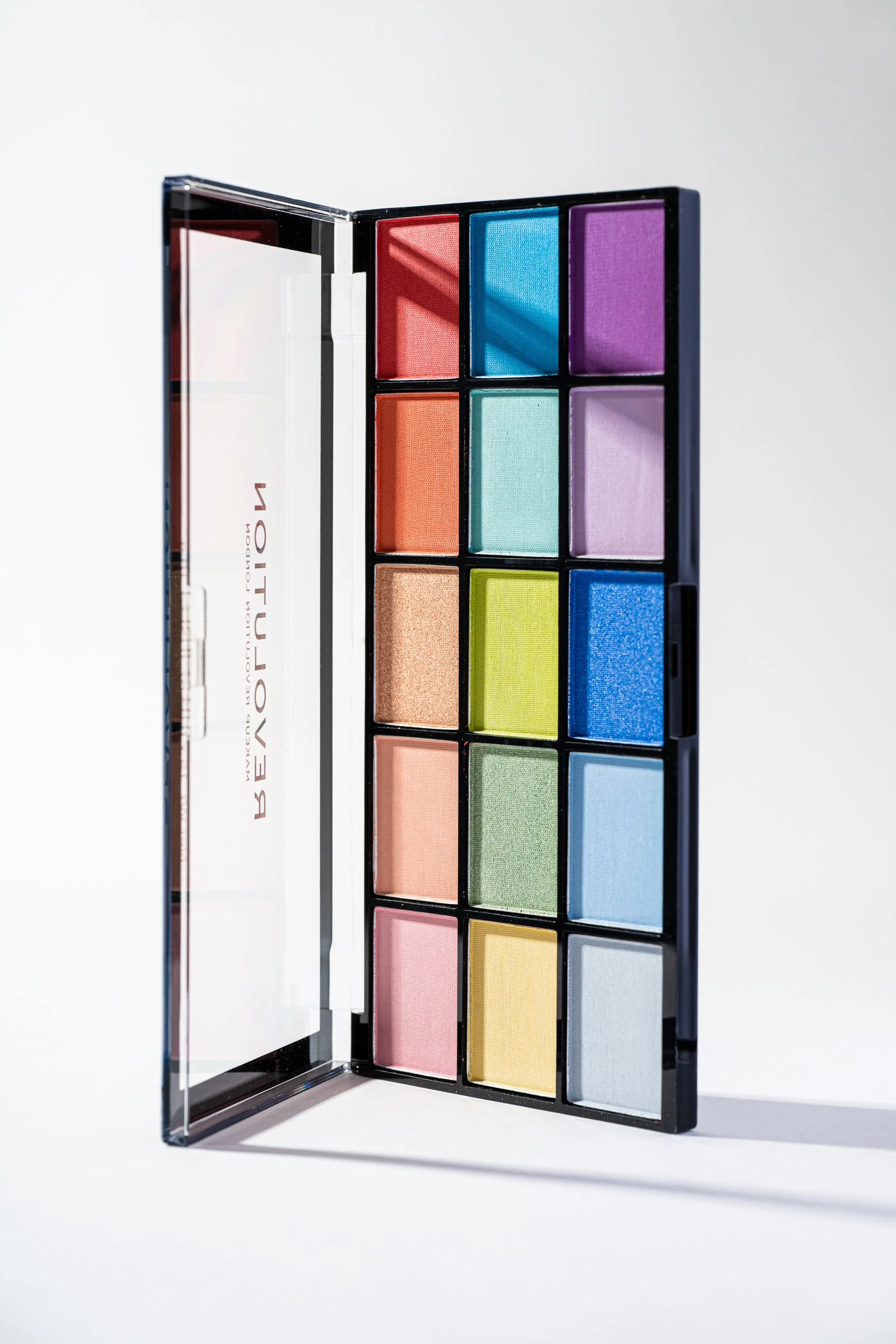 An open eyeshadow palette with multiple colorful eyeshadows arranged in a grid, set against a plain white background.
