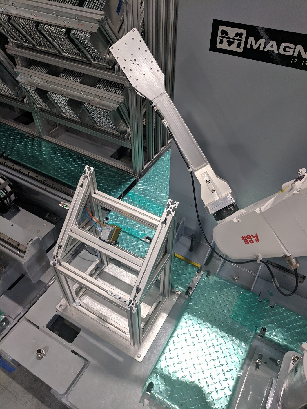 Industrial robotic arm assembling or handling a metallic frame in a manufacturing setting with safety and equipment components visible.