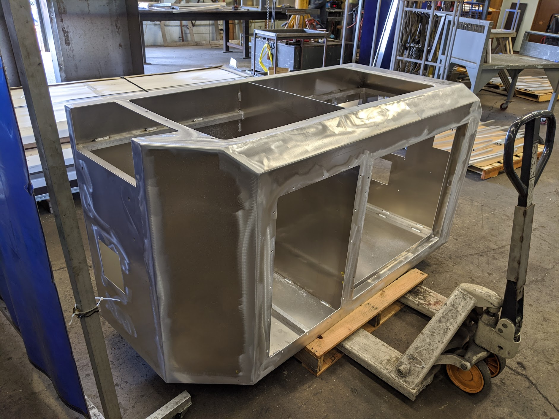 A metal frame structure under construction in a workshop, placed on a hand truck and a wooden pallet, with various tools and parts in the background.