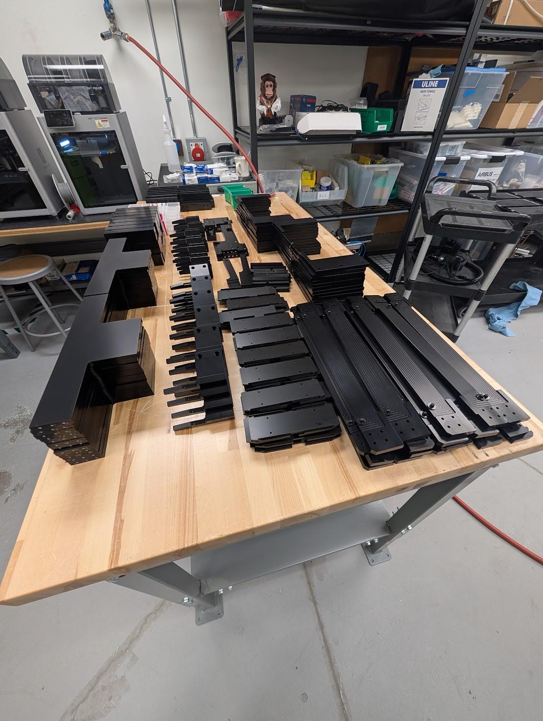 Array of black metal parts and components laid out on a wooden workbench in a workshop.