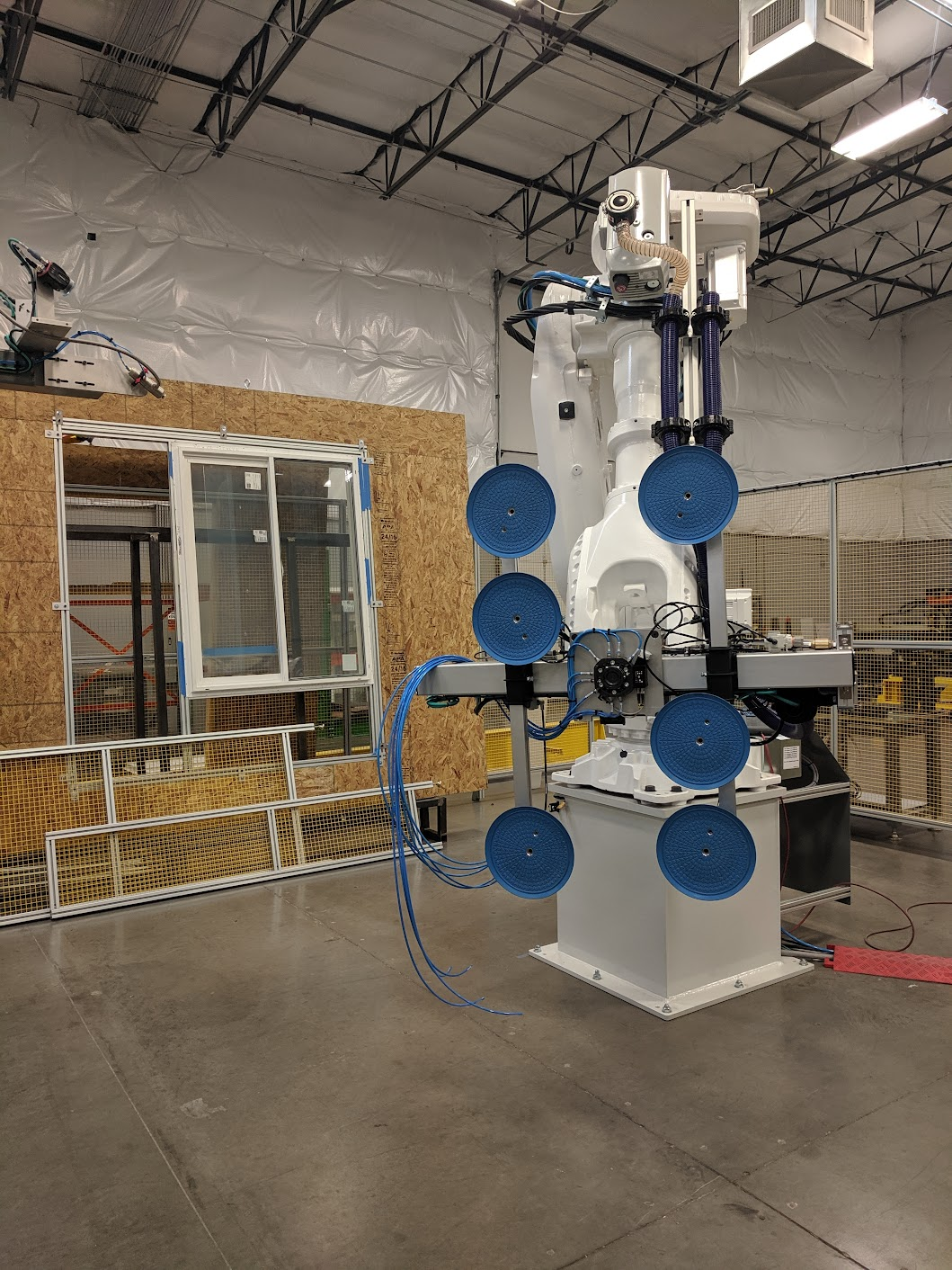A large industrial robotic arm with a white body and multiple blue circular pads is standing in a factory or lab environment, with exposed ceiling pipes and a plywood wall in the background.