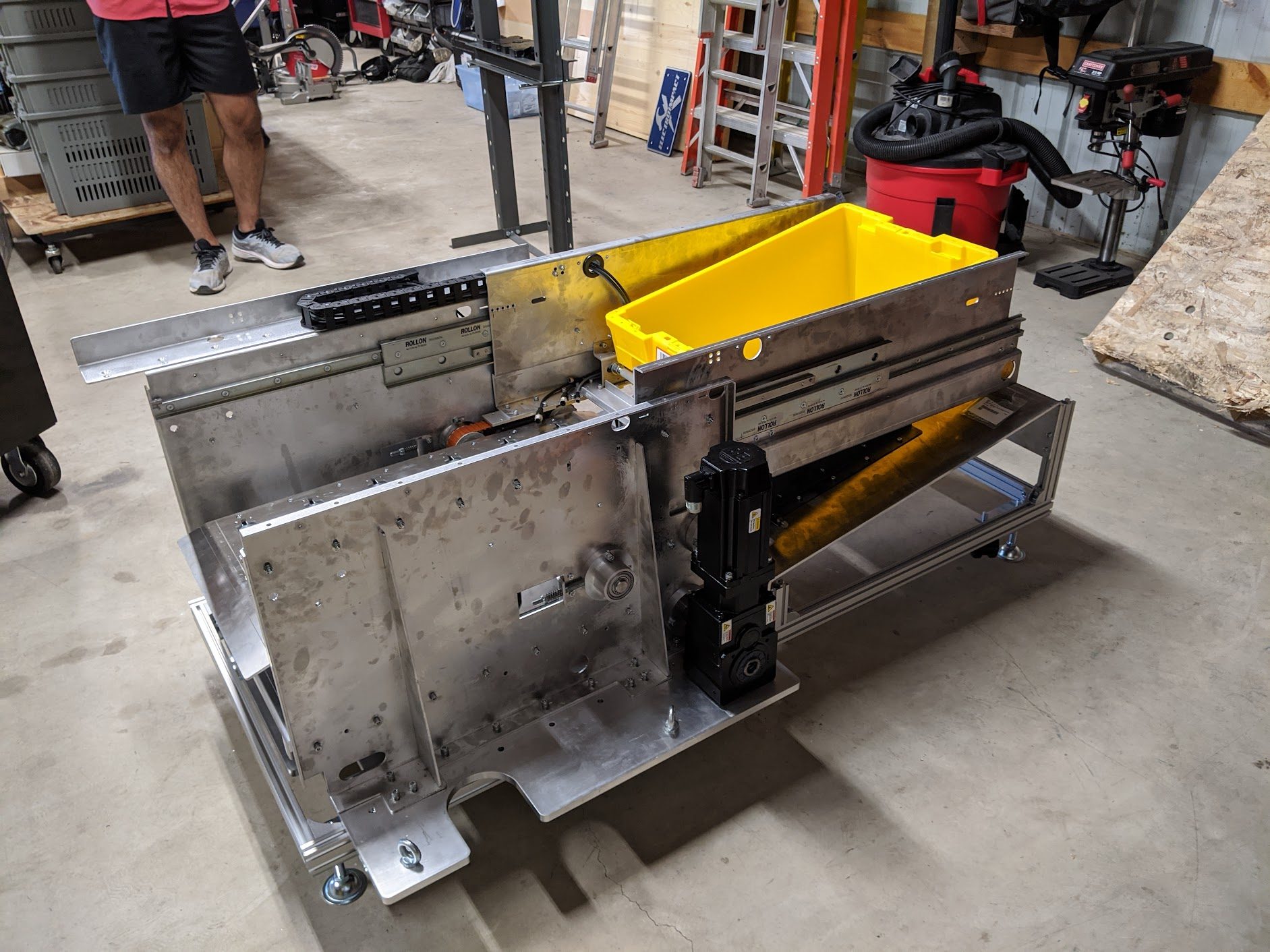 A robotic assembly machine made of metal with a yellow storage bin on top, situated in a workshop with tools and equipment in the background.