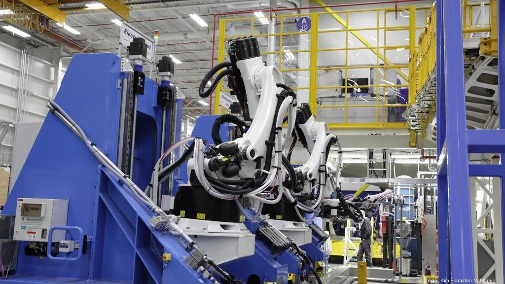Industrial factory setting with robotic arms working on machinery, with people in the background and safety railings.