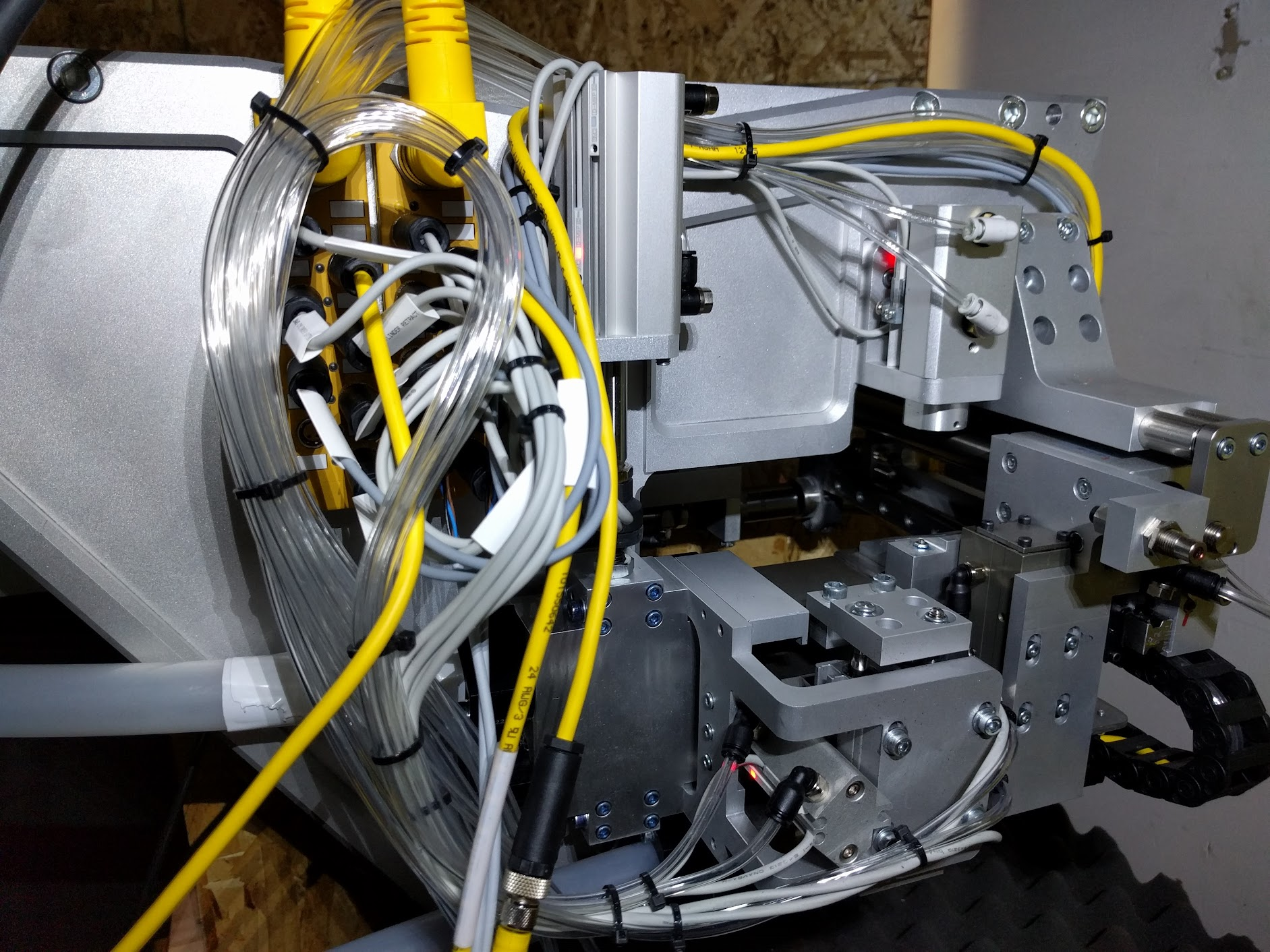 Close-up of a complex robotic or industrial machinery with metal parts, numerous cables, and tubing, including yellow, white, and transparent tubes, some bundled and secured with black ties.