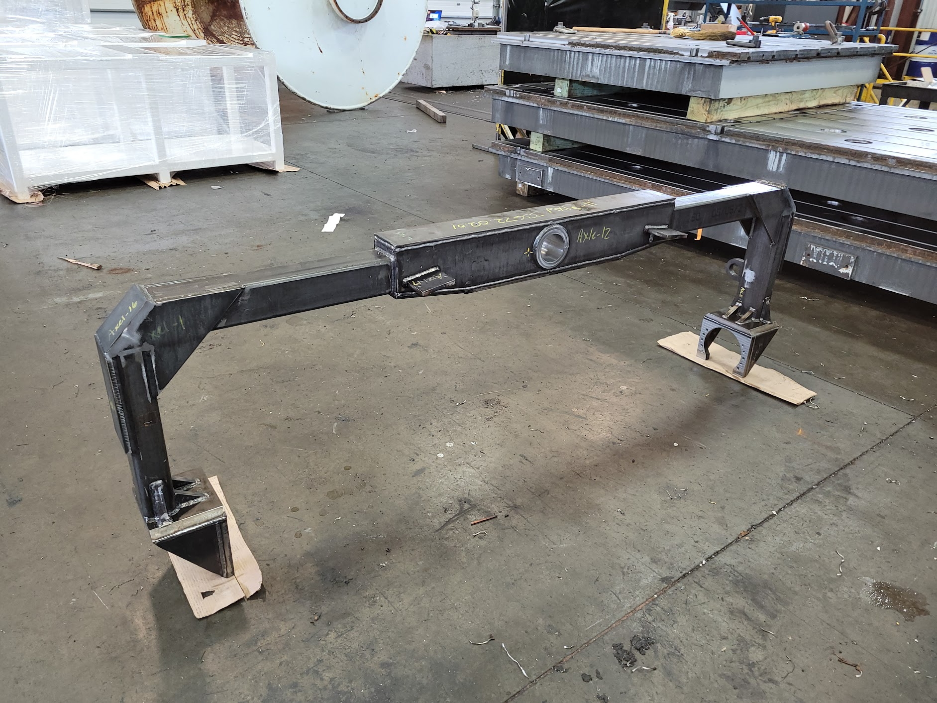 Metal framework or arm structure in an industrial workshop setting, with various metal parts, tools, and equipment in the background.