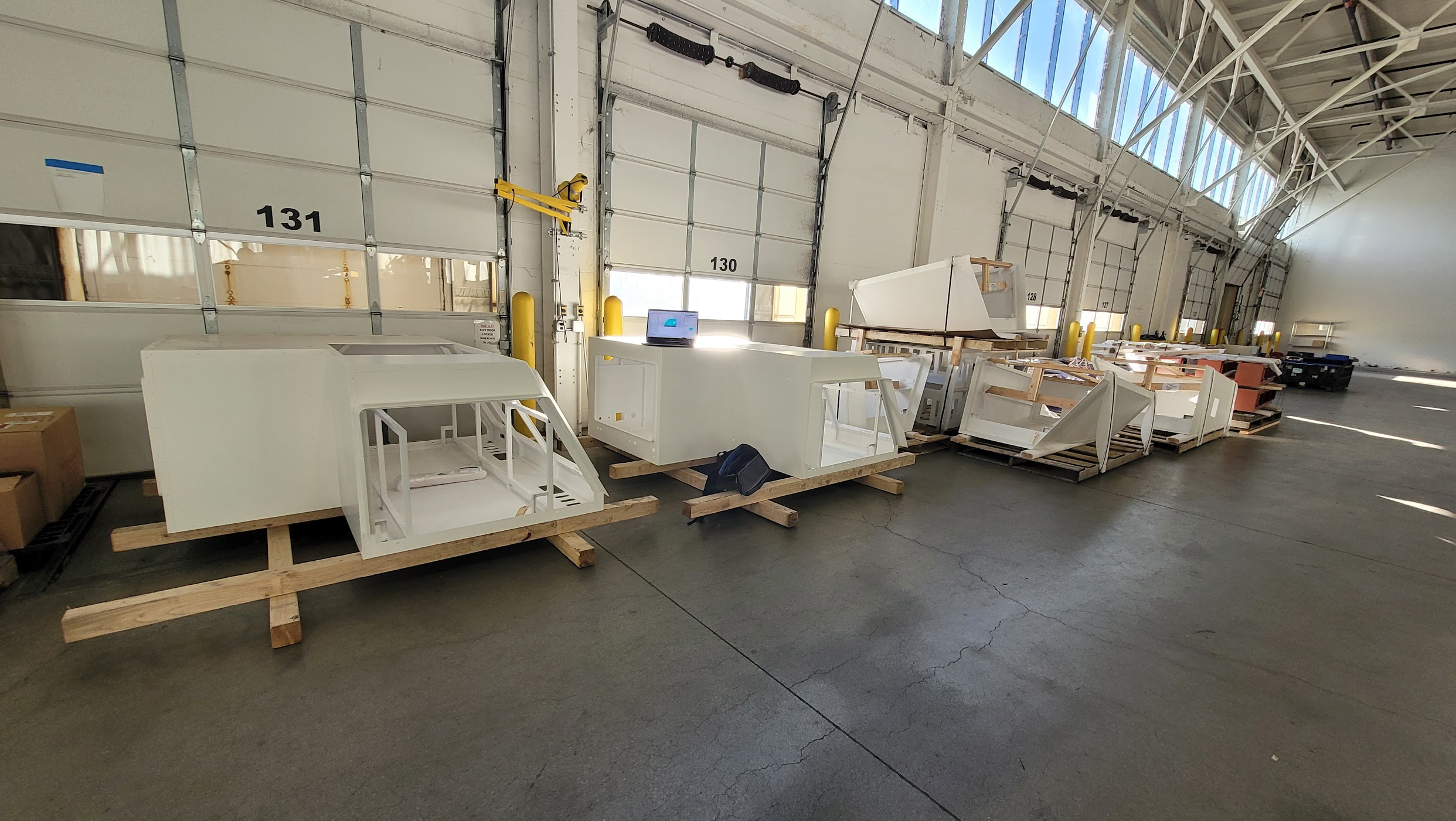 Several white refrigerator or freezer units are placed on wooden pallets inside a warehouse, with some rectangular packaging materials stacked nearby. The warehouse has high ceilings, large windows allowing natural light, and roll-up garage doors num