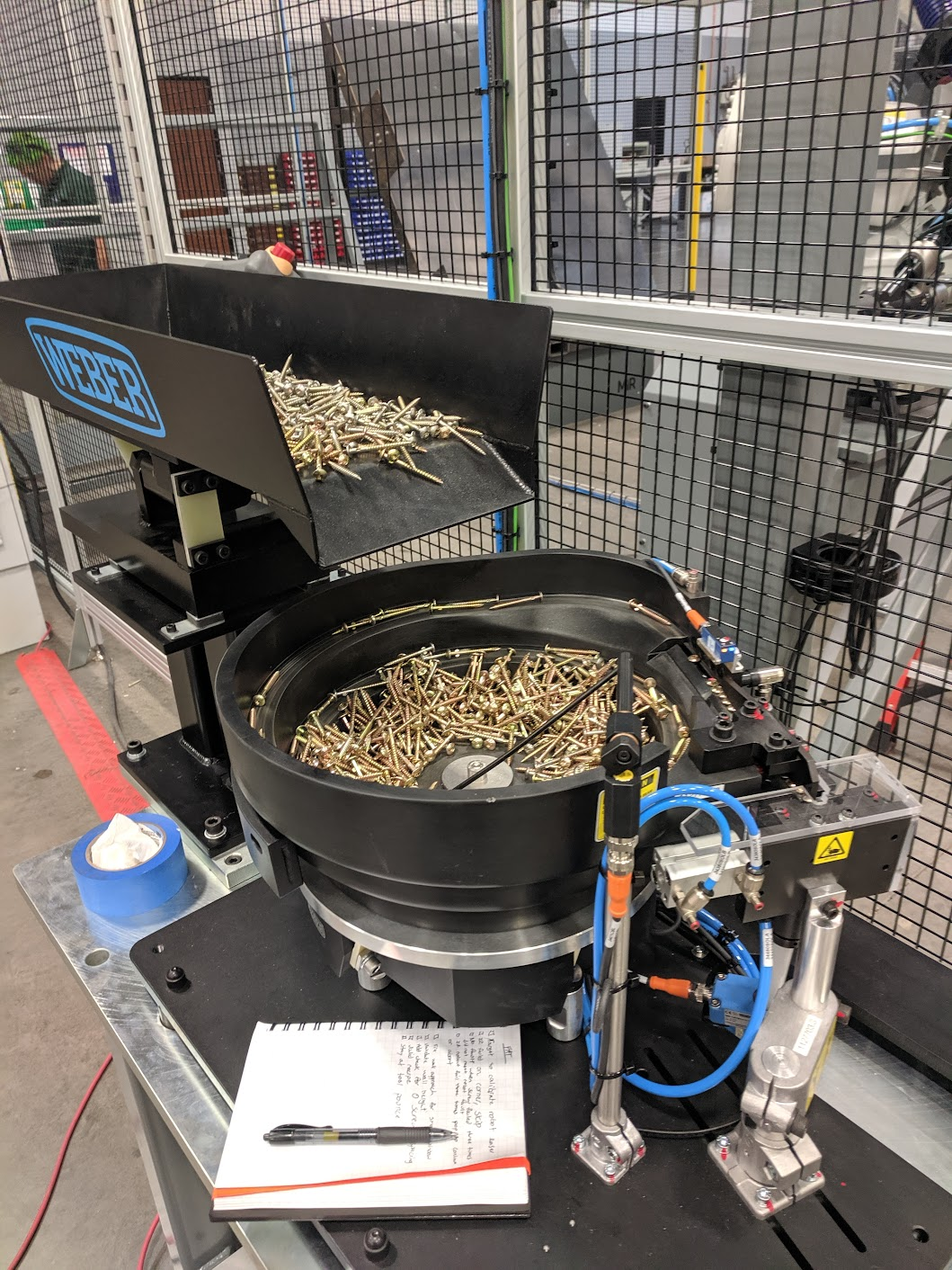 A mechanical device with a bowl containing gold-colored screws and a conveyor belt with additional screws, situated in an industrial setting with metal fencing, tools, and equipment in the background.