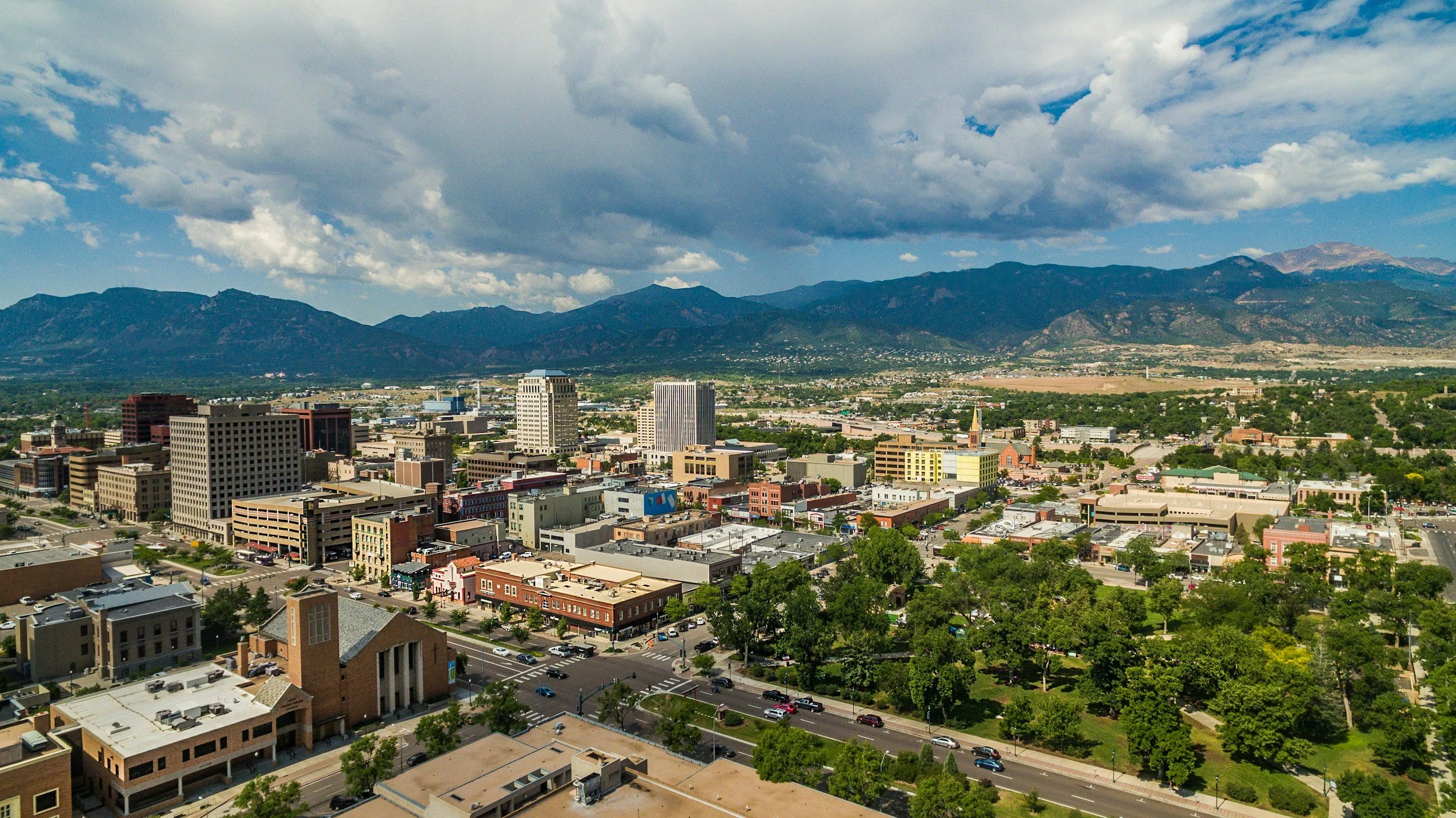 A cityscape with tall buildings, green trees, and a mountain range in the background under a partly cloudy sky.
