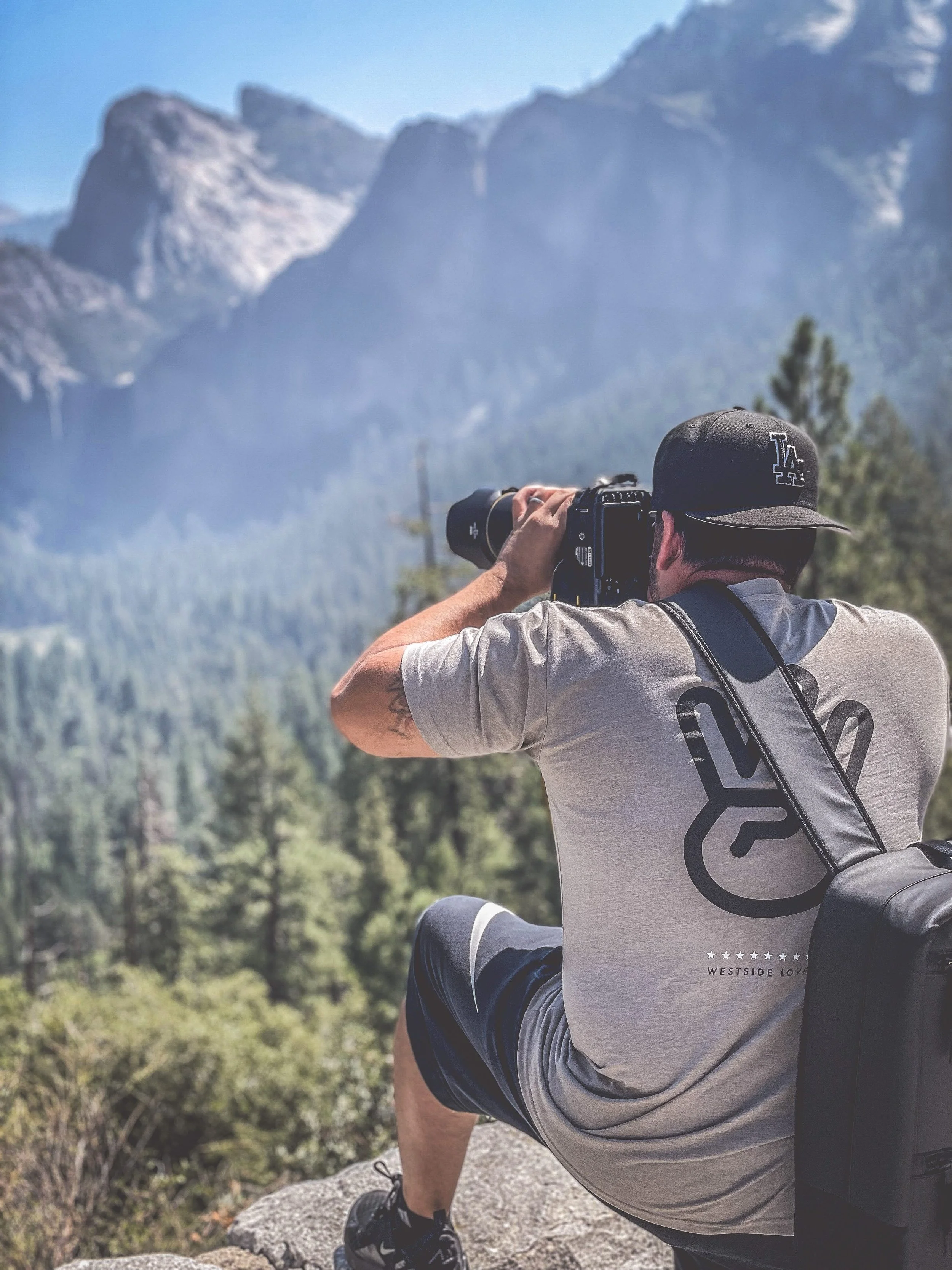 A man with a camera taking a photo of mountains and trees in a scenic outdoor location.