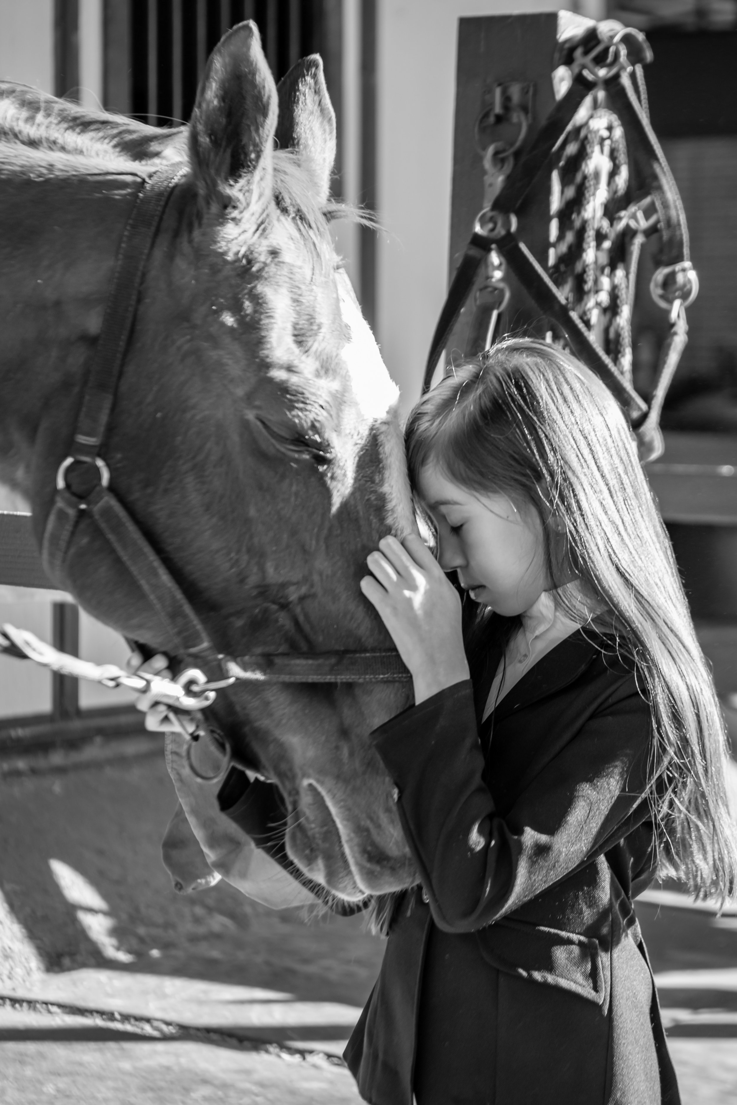 A young girl with long hair in a coat touches foreheads with a horse, both with closed eyes, in a black-and-white photo.