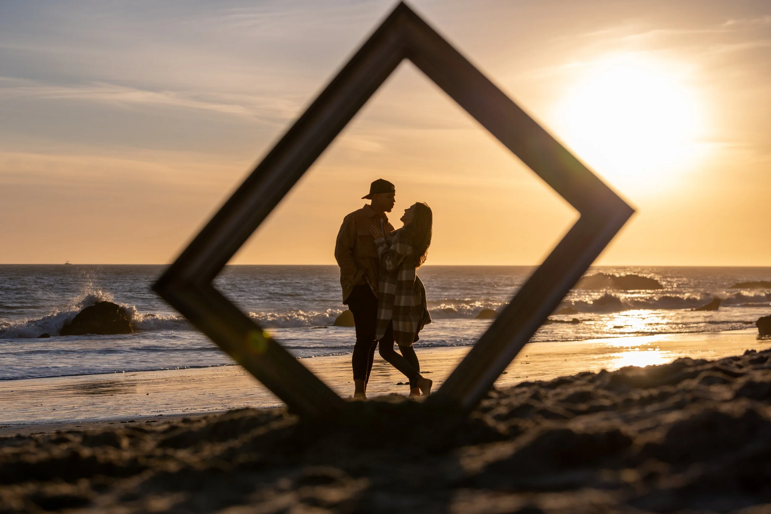 A couple standing on the beach during sunset, seen through a tilted picture frame.