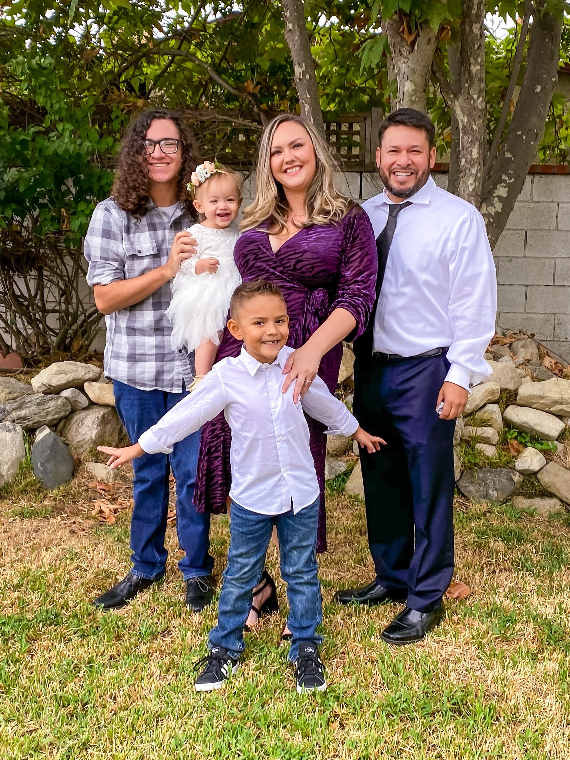 Family of five posing outdoors in a backyard with trees and rocks in the background. The group includes a woman in a purple dress, a man in a white shirt and black tie, a girl in a plaid shirt, a boy in a white shirt and jeans, and a young girl in a white dress holding a flower headband, all smiling.