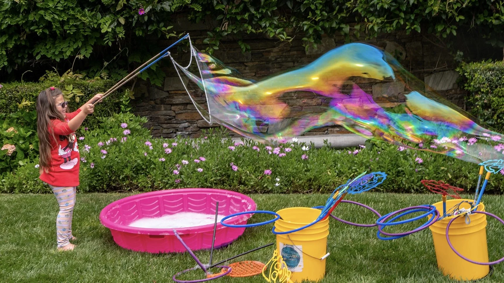 A young girl in a red shirt and striped pants is creating a giant soap bubble using a rope and sticks outdoors in a garden surrounded by green plants and pink flowers.