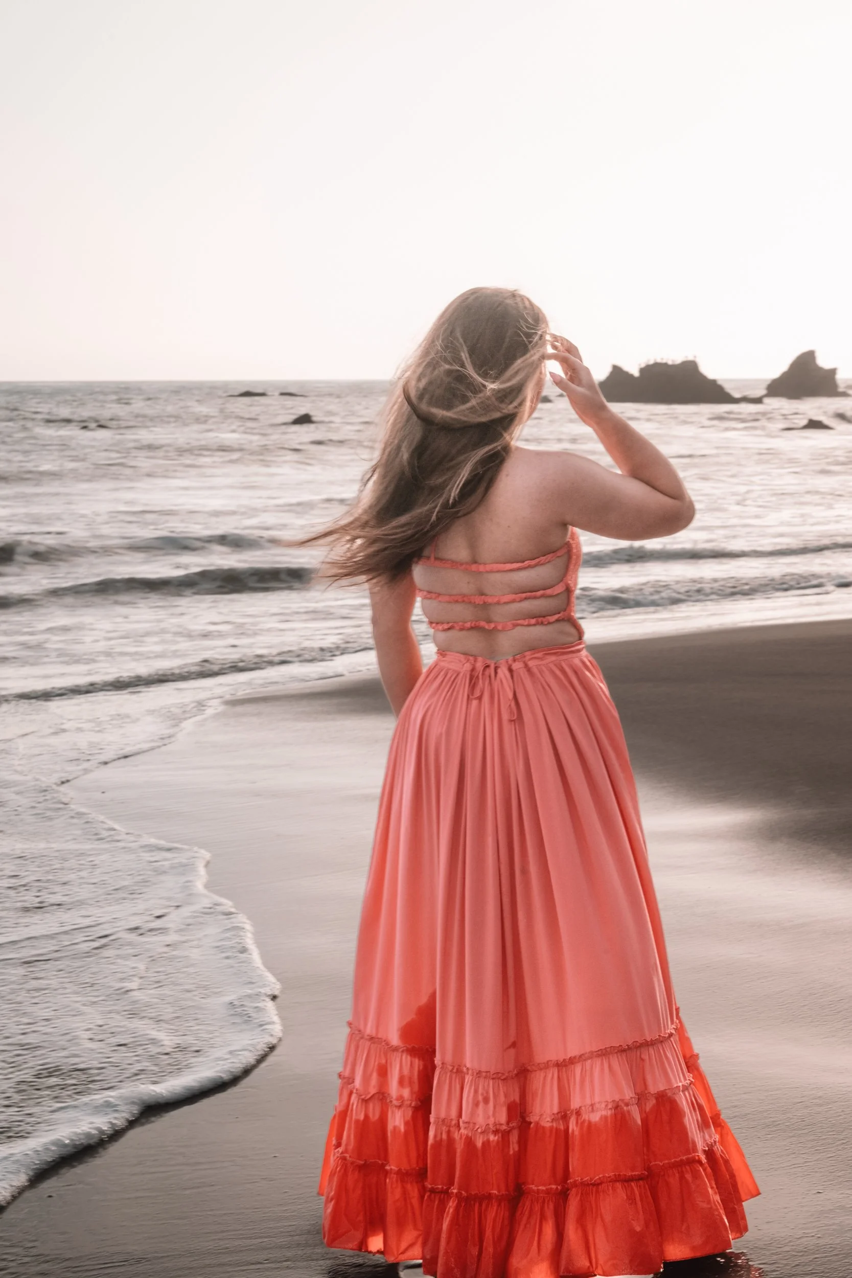 A woman with long hair standing on a beach, facing the ocean, wearing a coral-colored dress with ruffled layers.