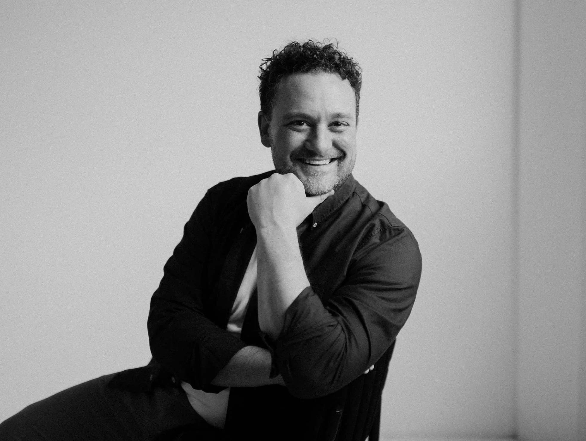 Black and white photo of a smiling man with curly hair, sitting and resting his chin on his hand, against a plain background.