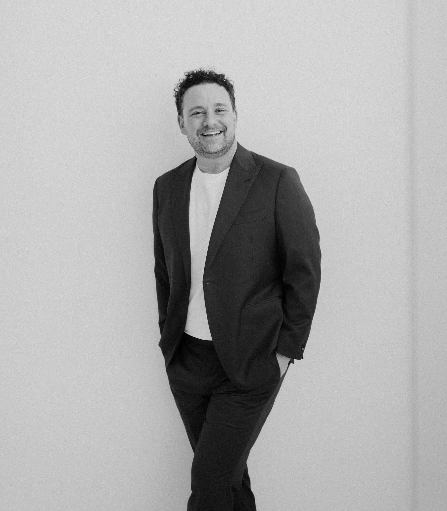 Man smiling in a black suit and white shirt standing against a plain wall.