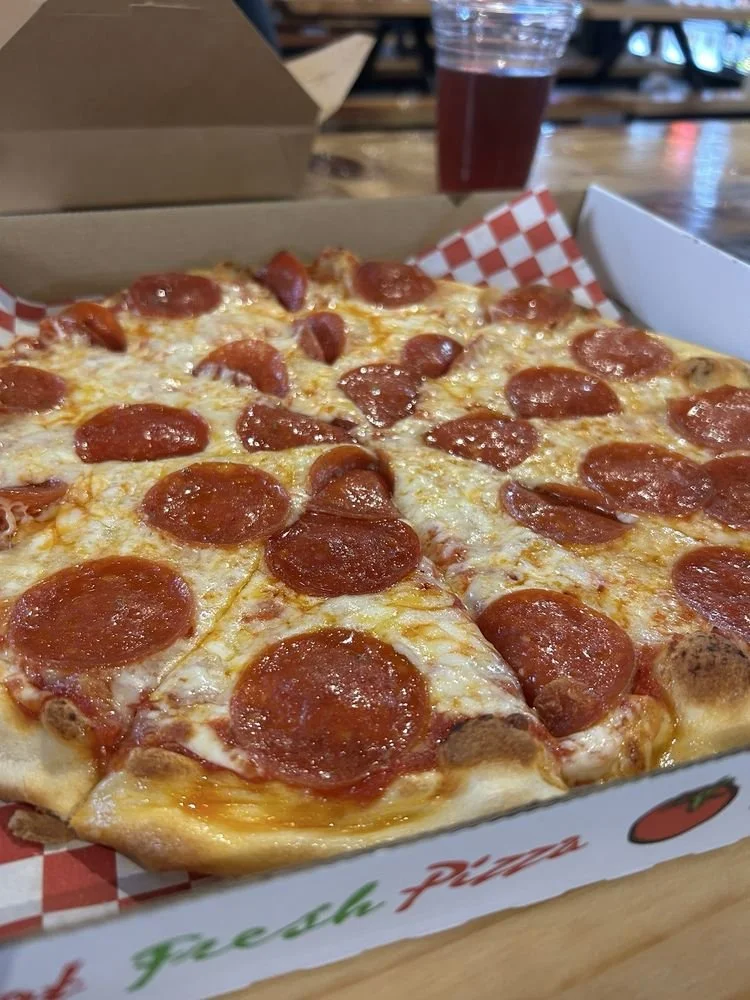 A pizza with pepperoni slices in a box on a wooden table, with a drink in the background.