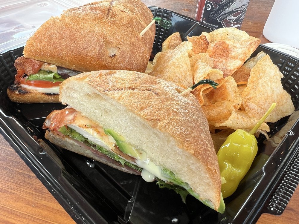 A black plastic takeout container with a sandwich on a bun, potato chips, and pepperoncini peppers.