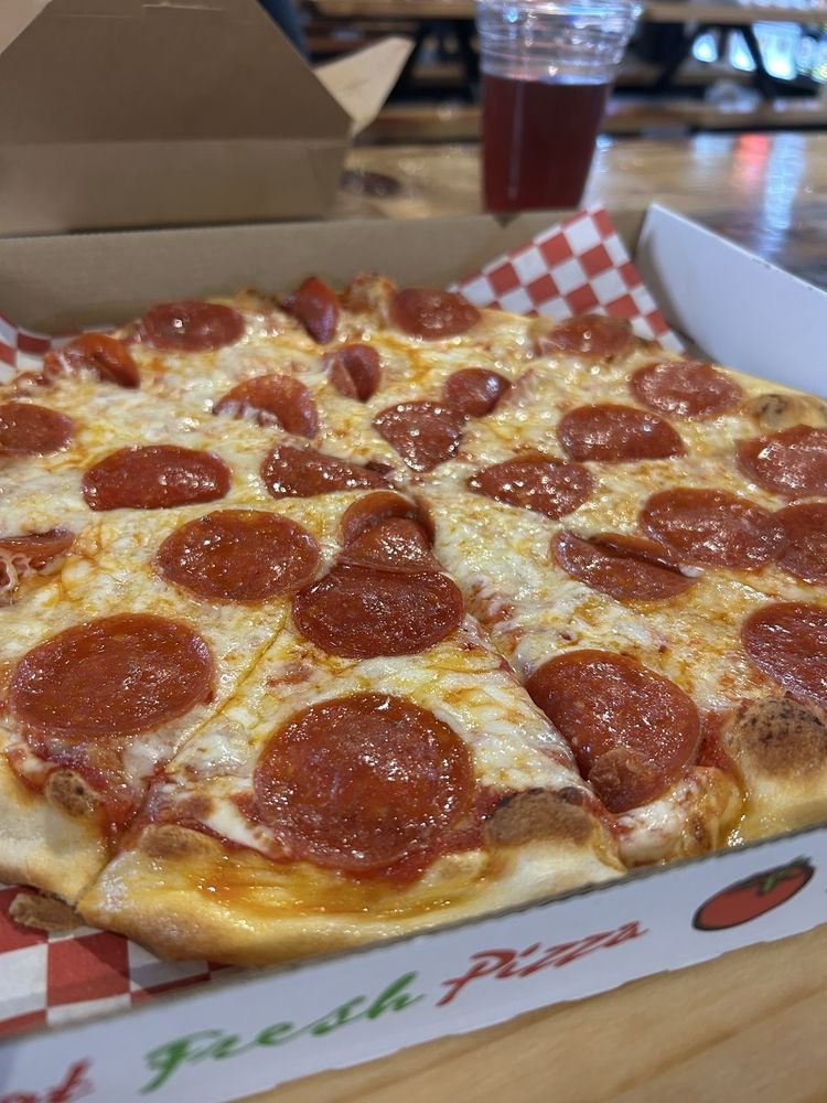 A pepperoni pizza in an open pizza box with a checkerboard liner, in a restaurant setting with a glass of dark soda and wooden table in the background.