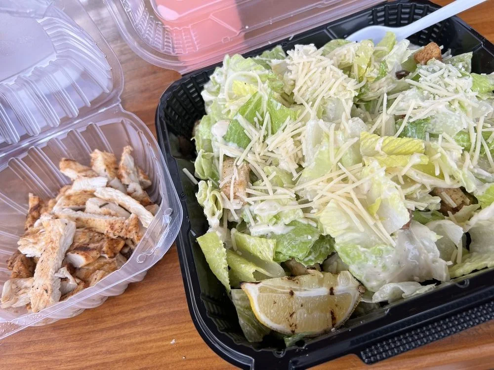 Caesar salad in a black takeout container with shredded cheese and lemon wedge, grilled chicken pieces in a separate clear plastic container, and pink salad dressing in a clear plastic container.