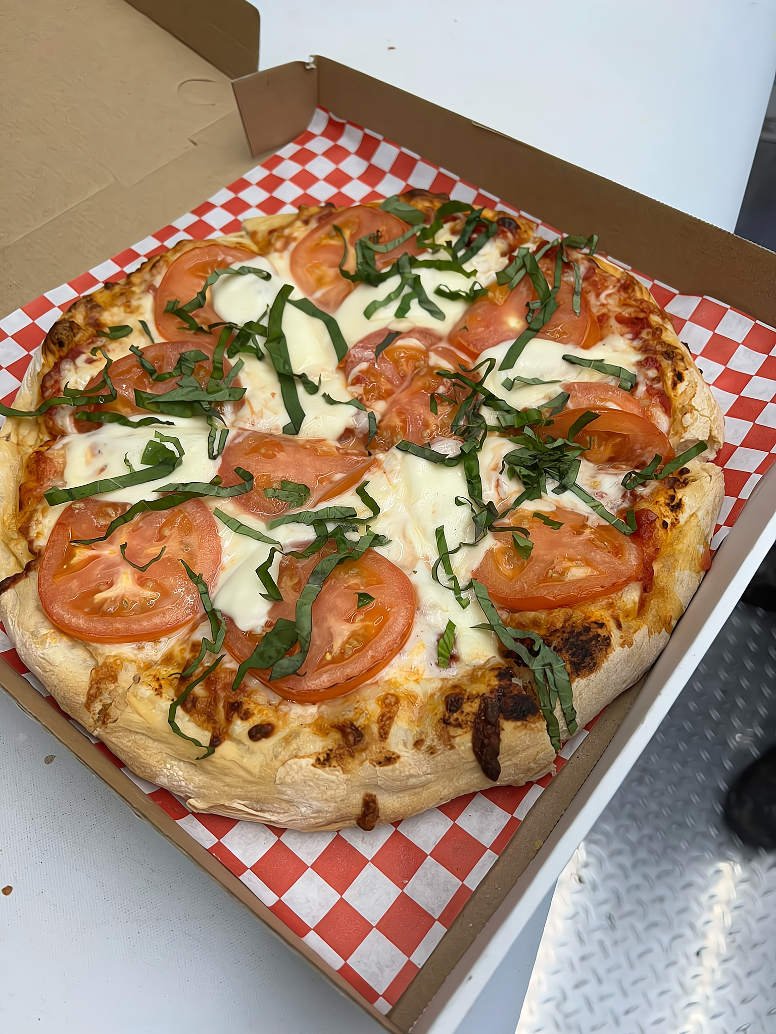 Pizza with tomato slices, mozzarella cheese, and basil on a checkered paper in a pizza box.